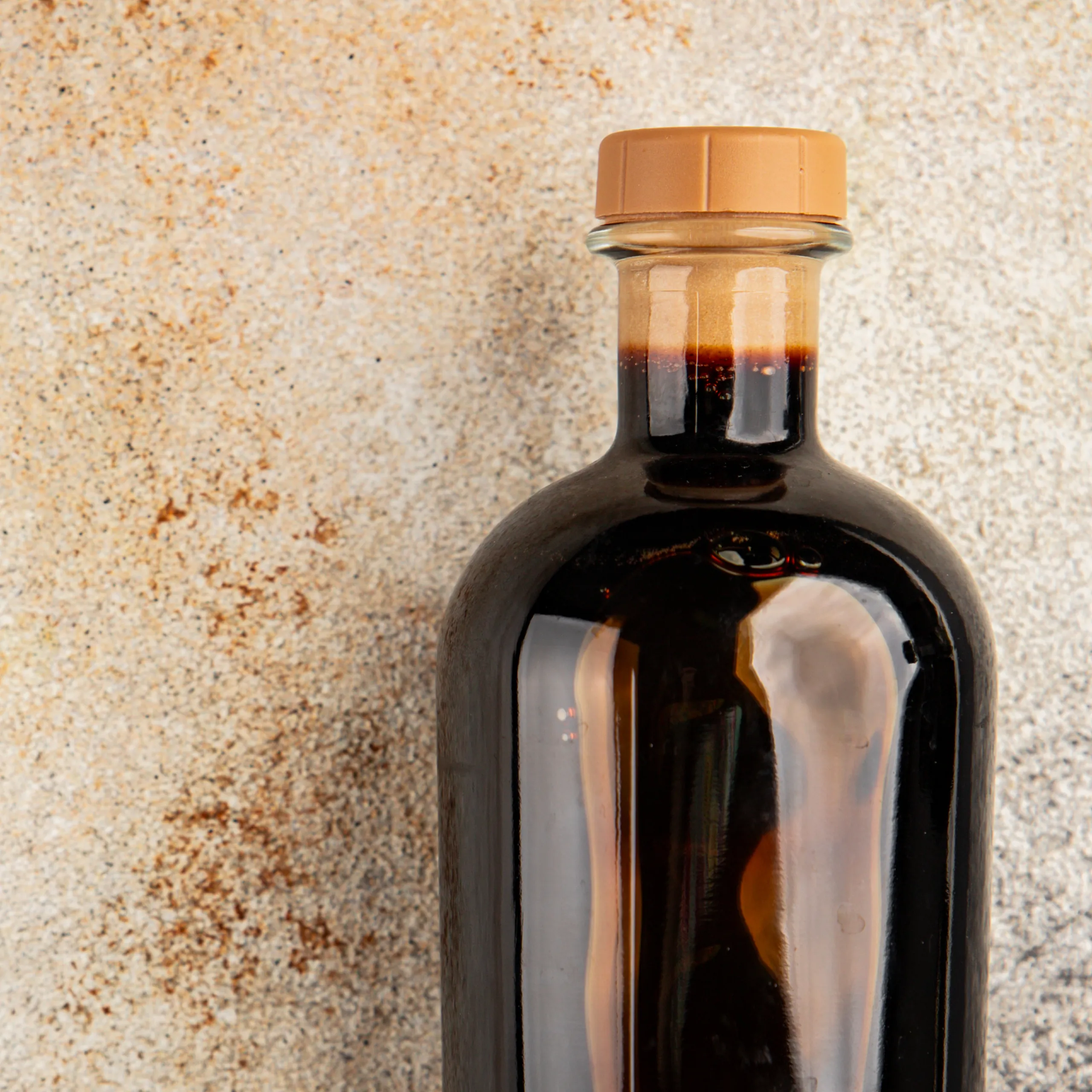 Close-up of a dark amber bottle neck with a light brown cap, against a speckled beige surface.