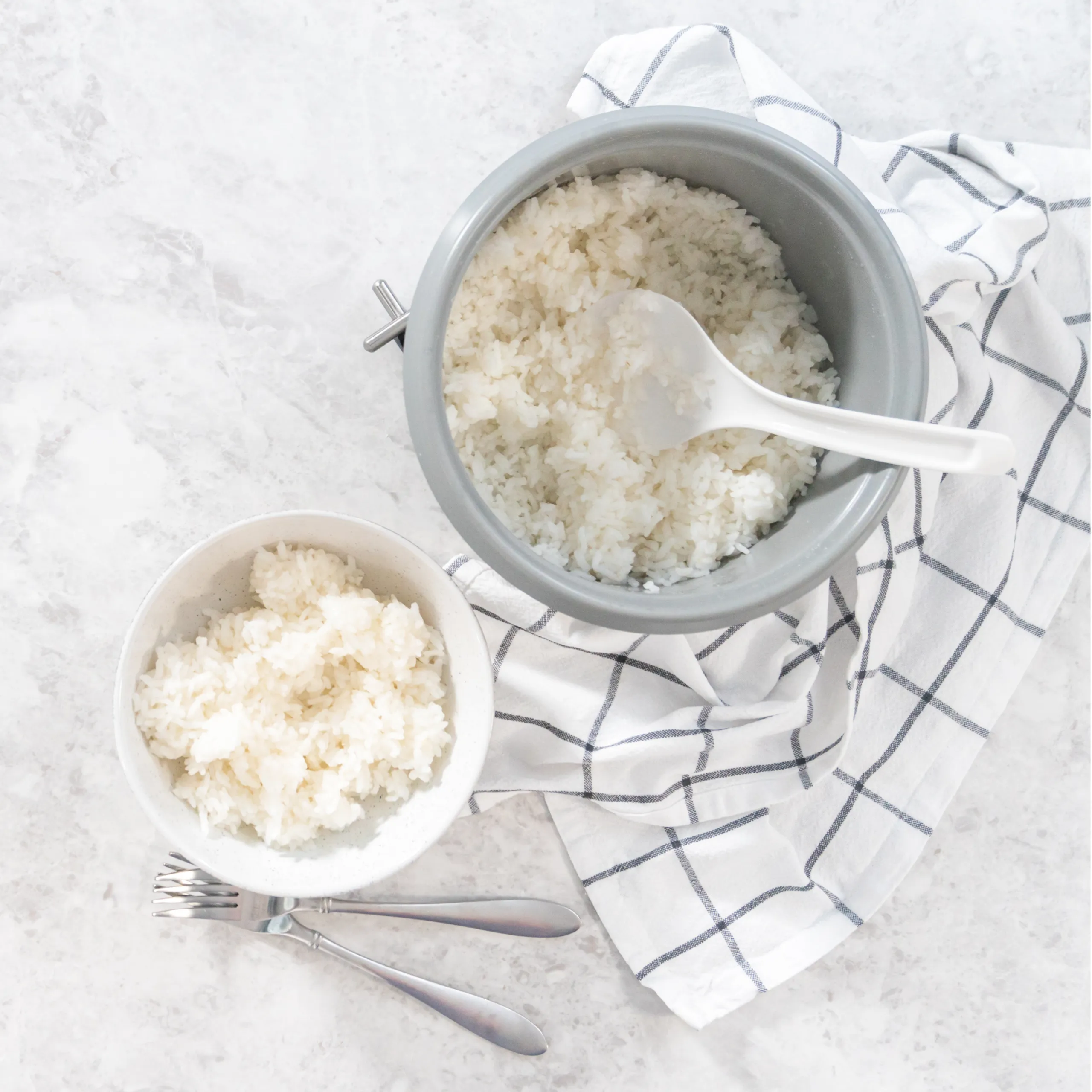 Two bowls of cooked white rice on a marble surface, with a white serving spoon and a striped cloth nearby.