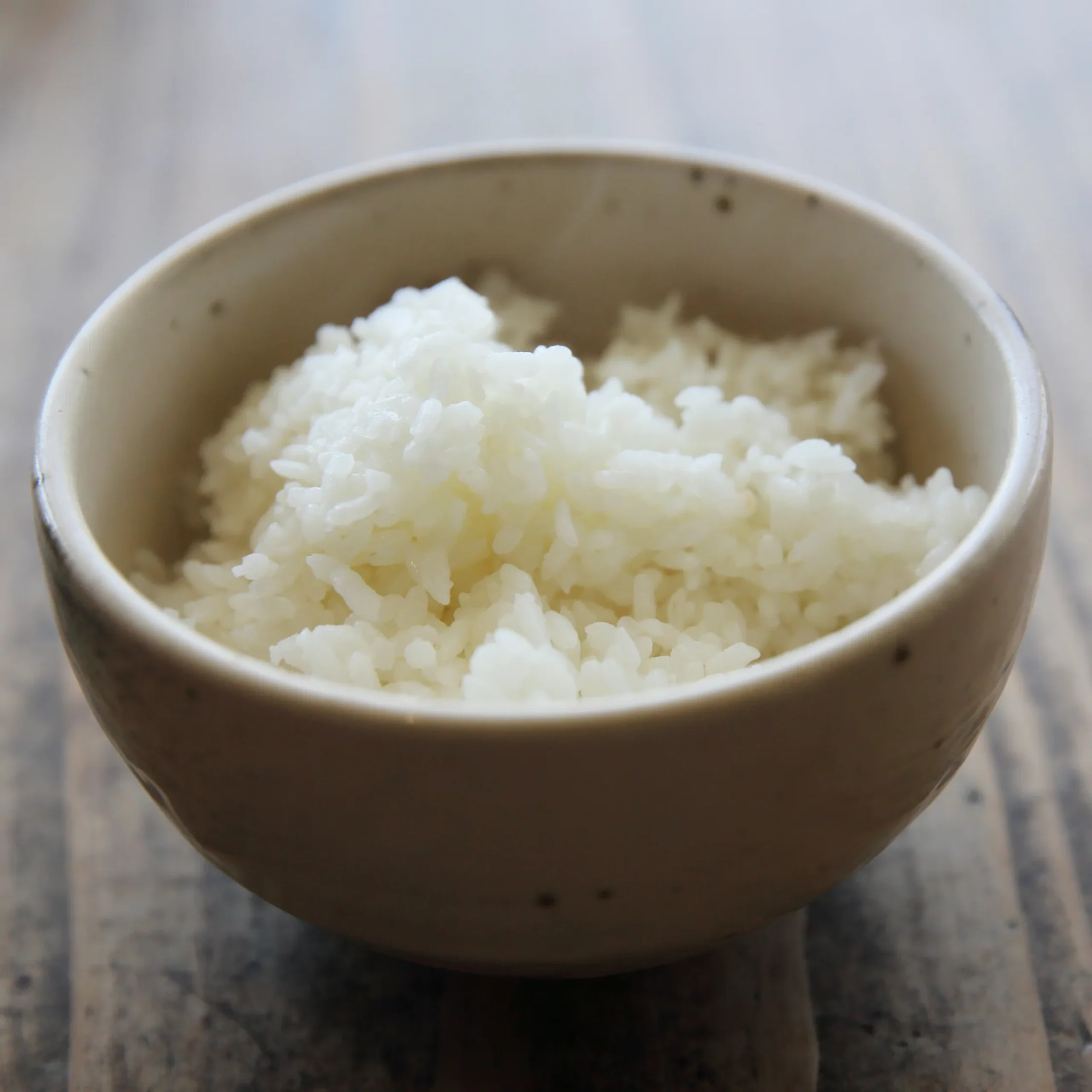 Close-up of a ceramic bowl filled with white rice on a wooden table, viewed from above.
