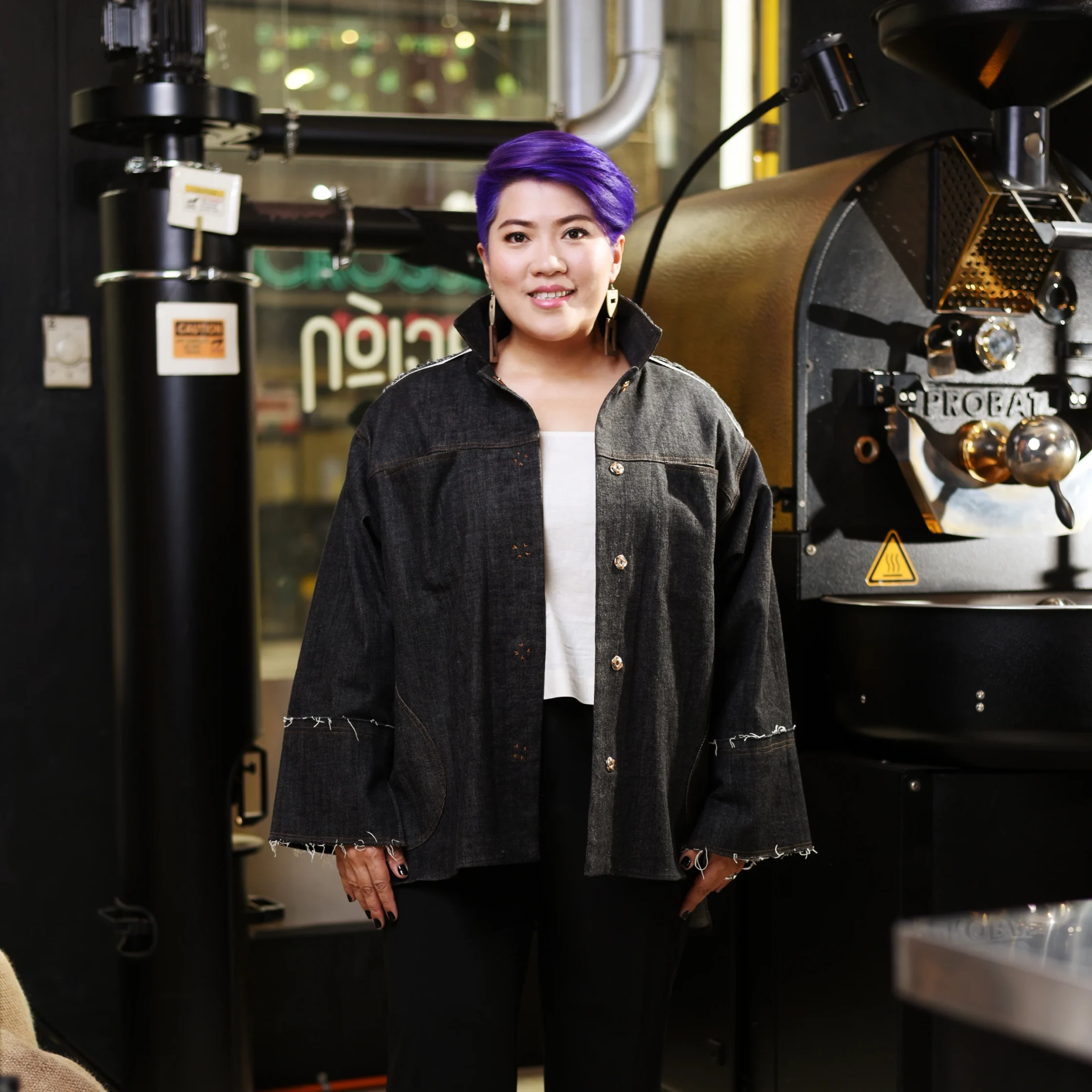 Person with short purple hair smiling, standing in front of large industrial machinery in a workshop setting.