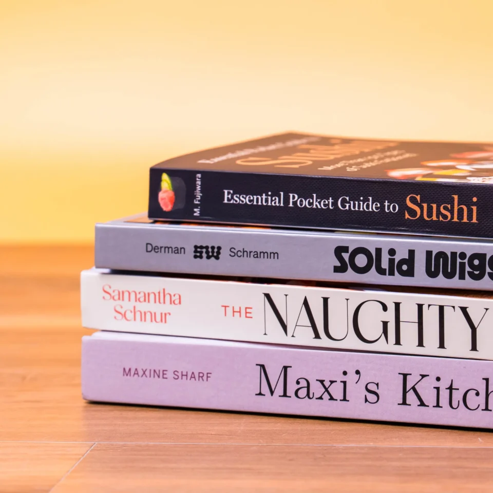 Stack of cookbooks stacked on a wooden floor; top book reads 'Essential Pocket Guide to Sushi' with other titles partially visible.