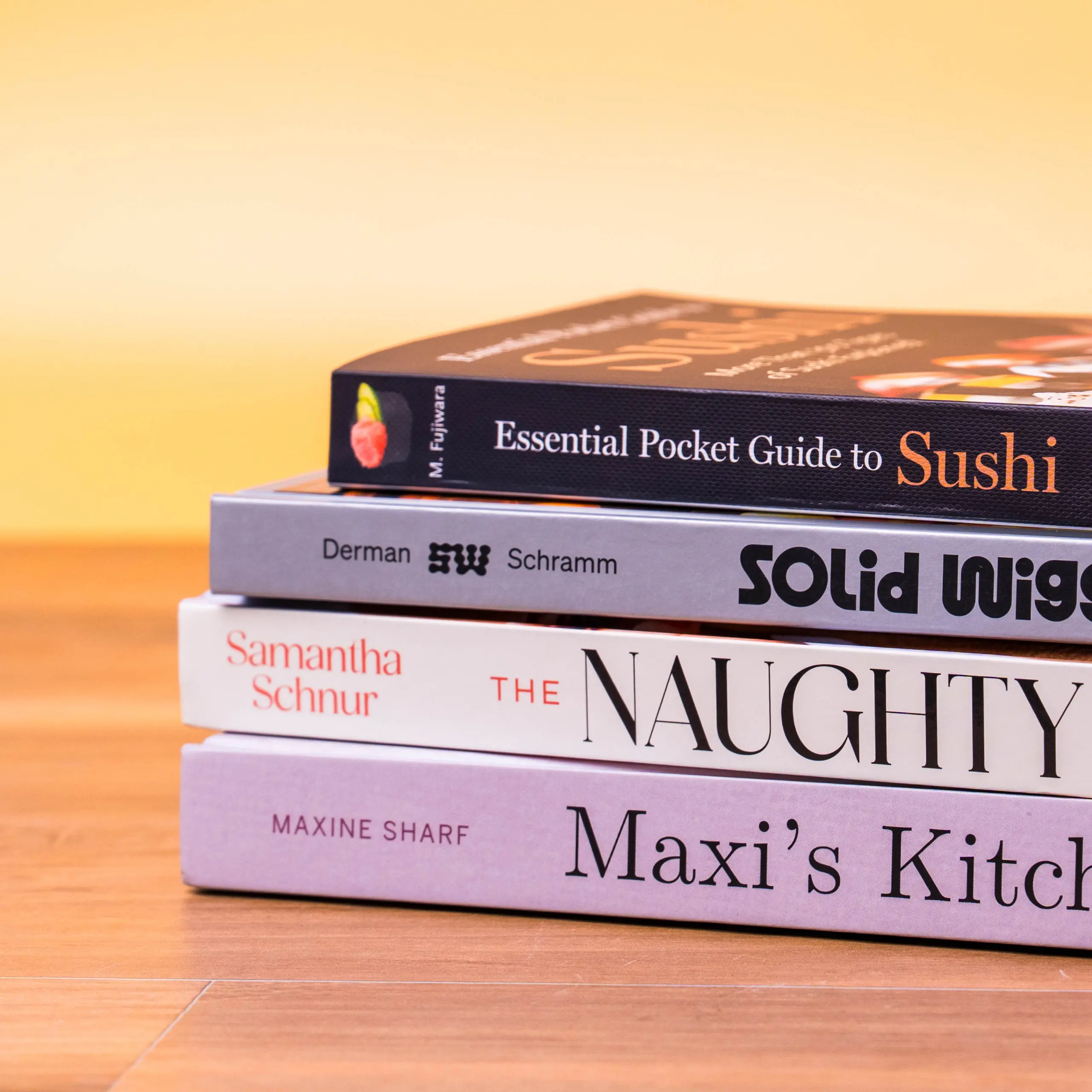 Stack of cookbooks stacked on a wooden floor; top book reads 'Essential Pocket Guide to Sushi' with other titles partially visible.