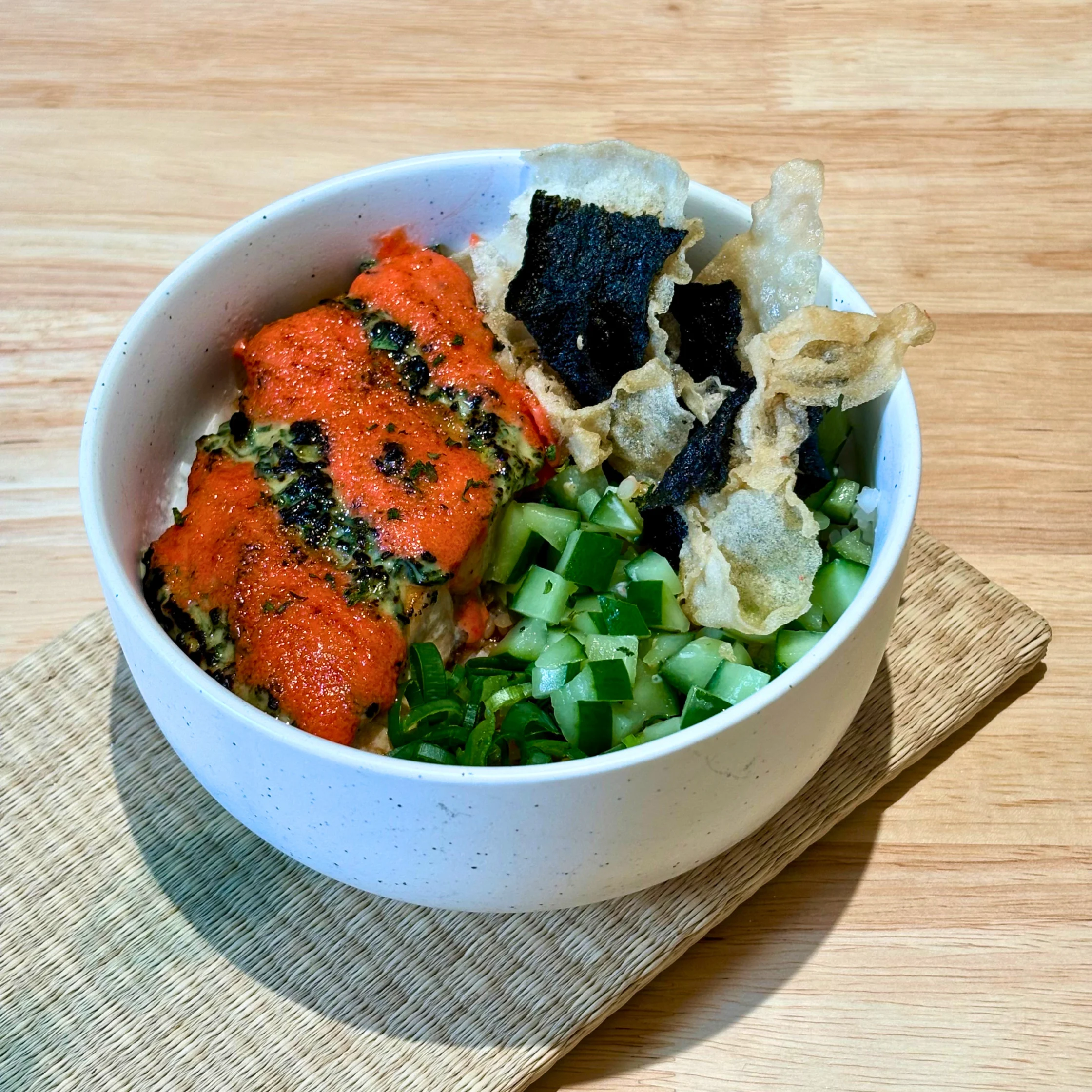 Close-up of a poke bowl with bright red spicy sauce, crispy seaweed chips, and chopped cucumber on a wooden surface.
