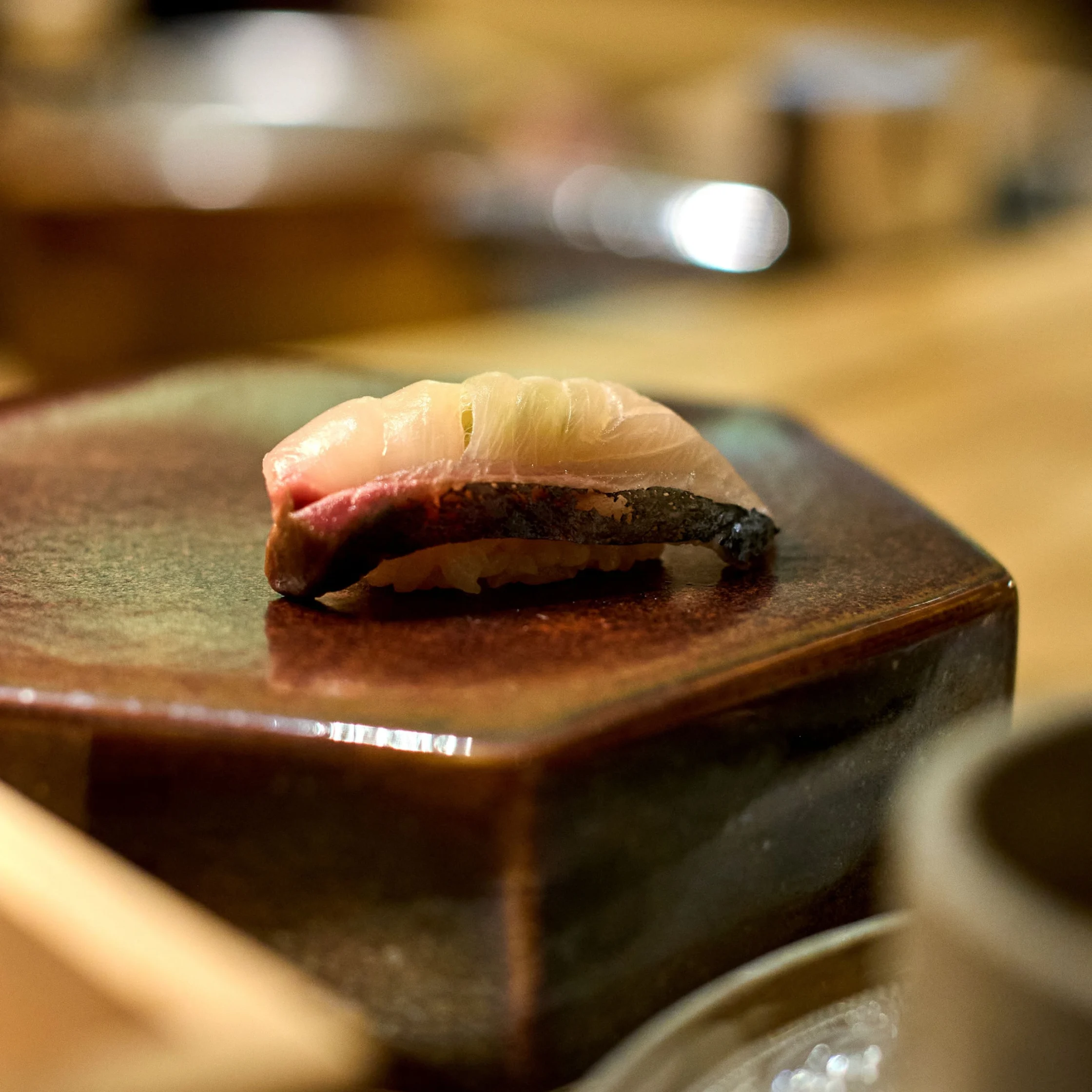 Out-of-focus close-up of a nigiri sushi piece resting on a dark plate under warm kitchen lighting.