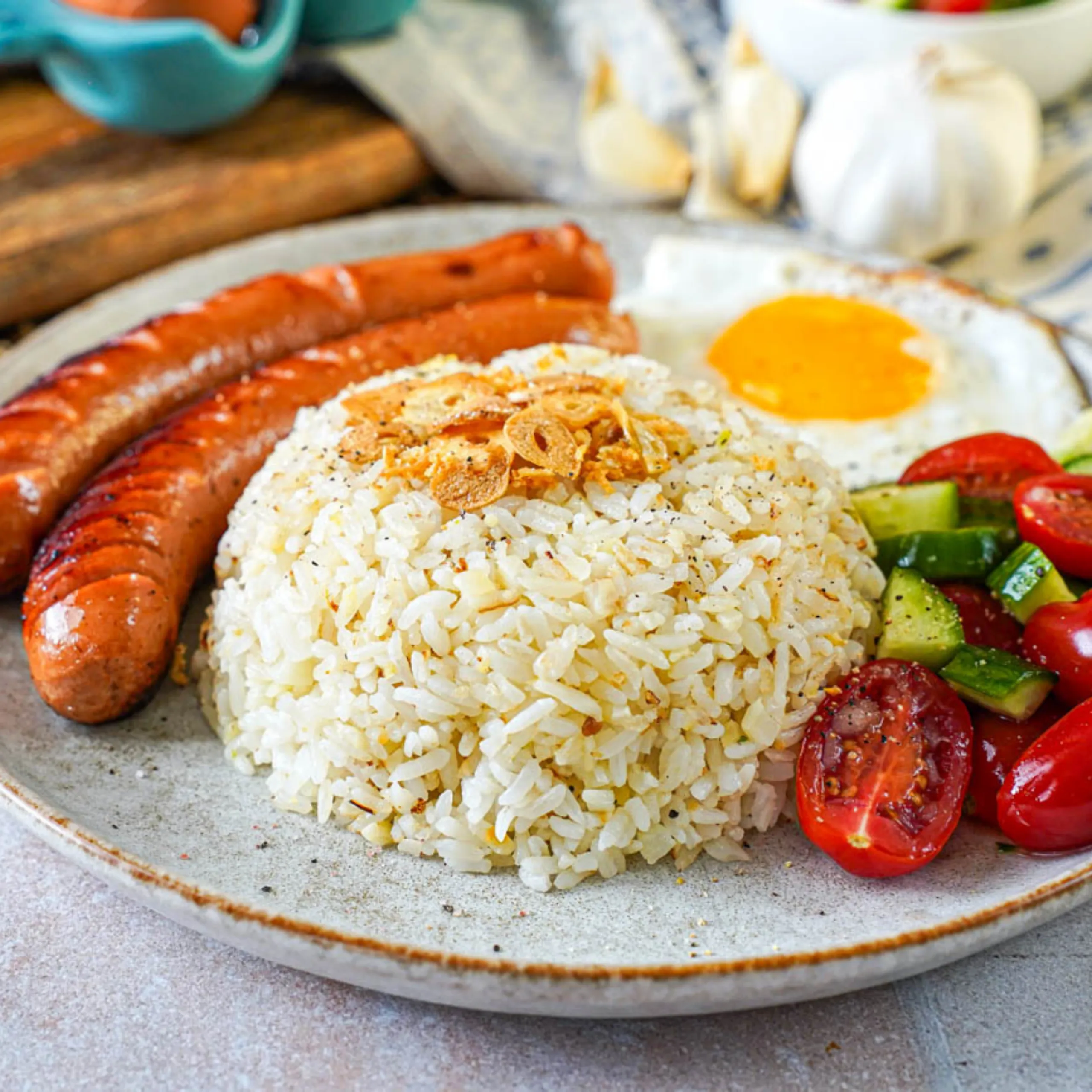 a plate with two long pieces of sausage, one sunny side up egg, sliced tomate, and a cup garlic fried rice