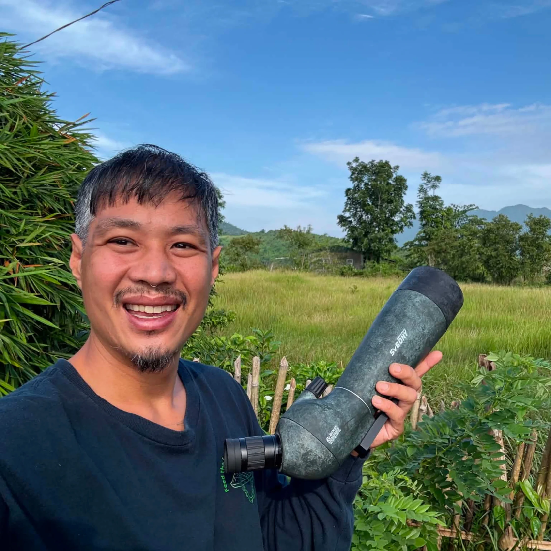 Smiling man taking a selfie outdoors with blue sky, fields, and trees in the background, holding a large gray cylindrical object. Regarded as a casual outdoor photo in a rural setting.