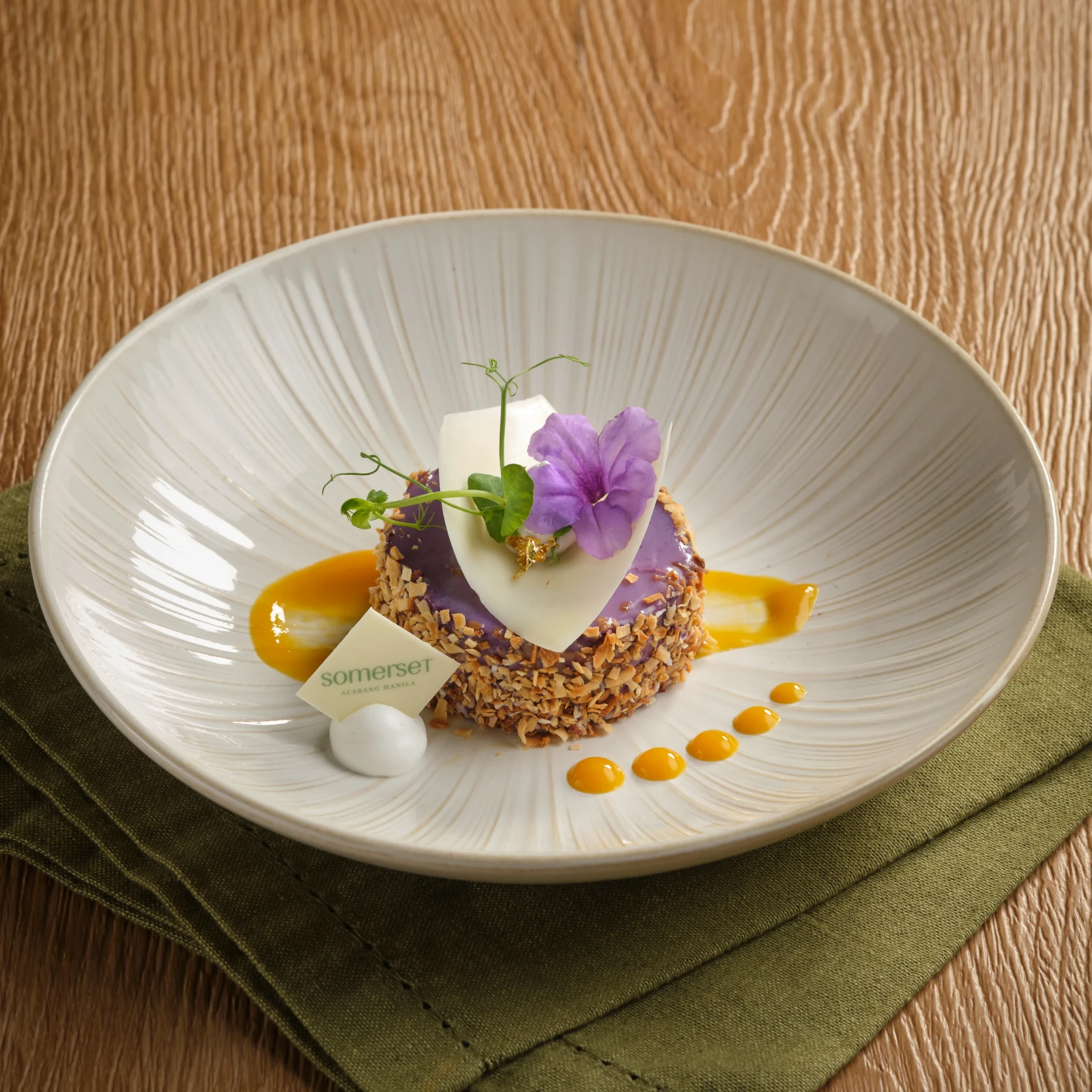 Dessert plated with purple glaze, white chocolate shard, crushed nuts, a purple flower garnish, and orange sauce dots on a white dish.
