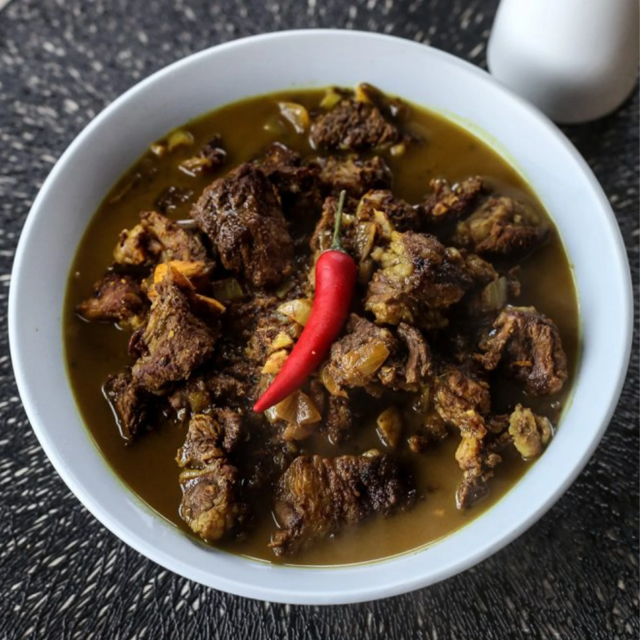 Beef curry with tender meat chunks in brown gravy, garnished with a red chili, served in a white bowl on a dark countertop.