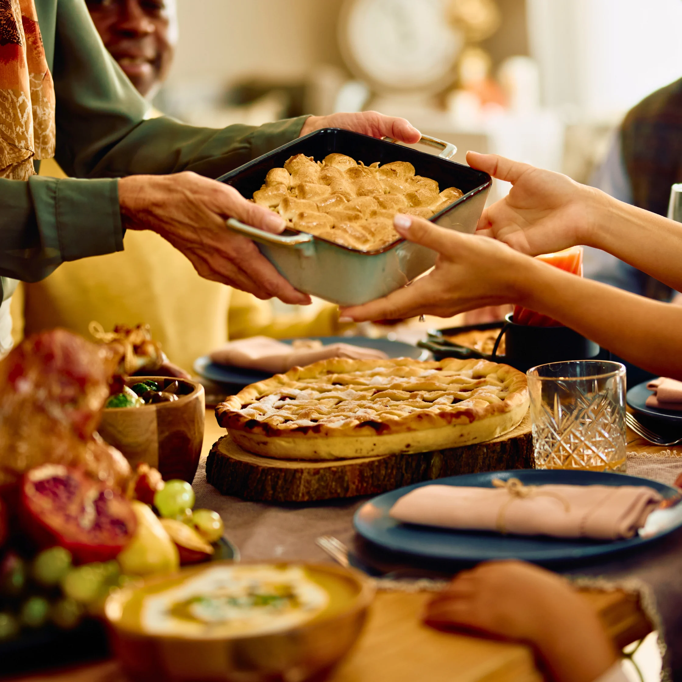 Hands passing a metal baking dish filled with a golden baked casserole to a guest at a table of pies.