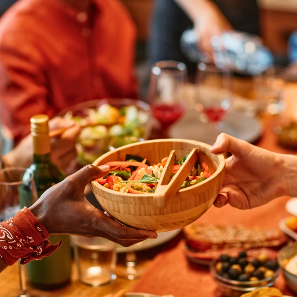Hands passing a wooden bowl of salad at a busy table during a meal.