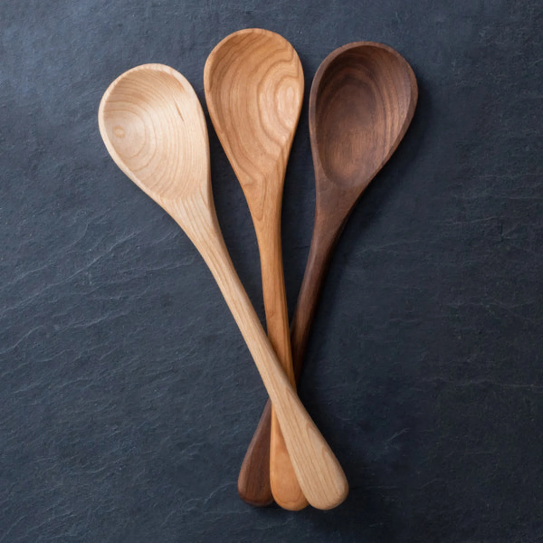 Three wooden spoons in light, medium, and dark wood arranged on a dark slate background.