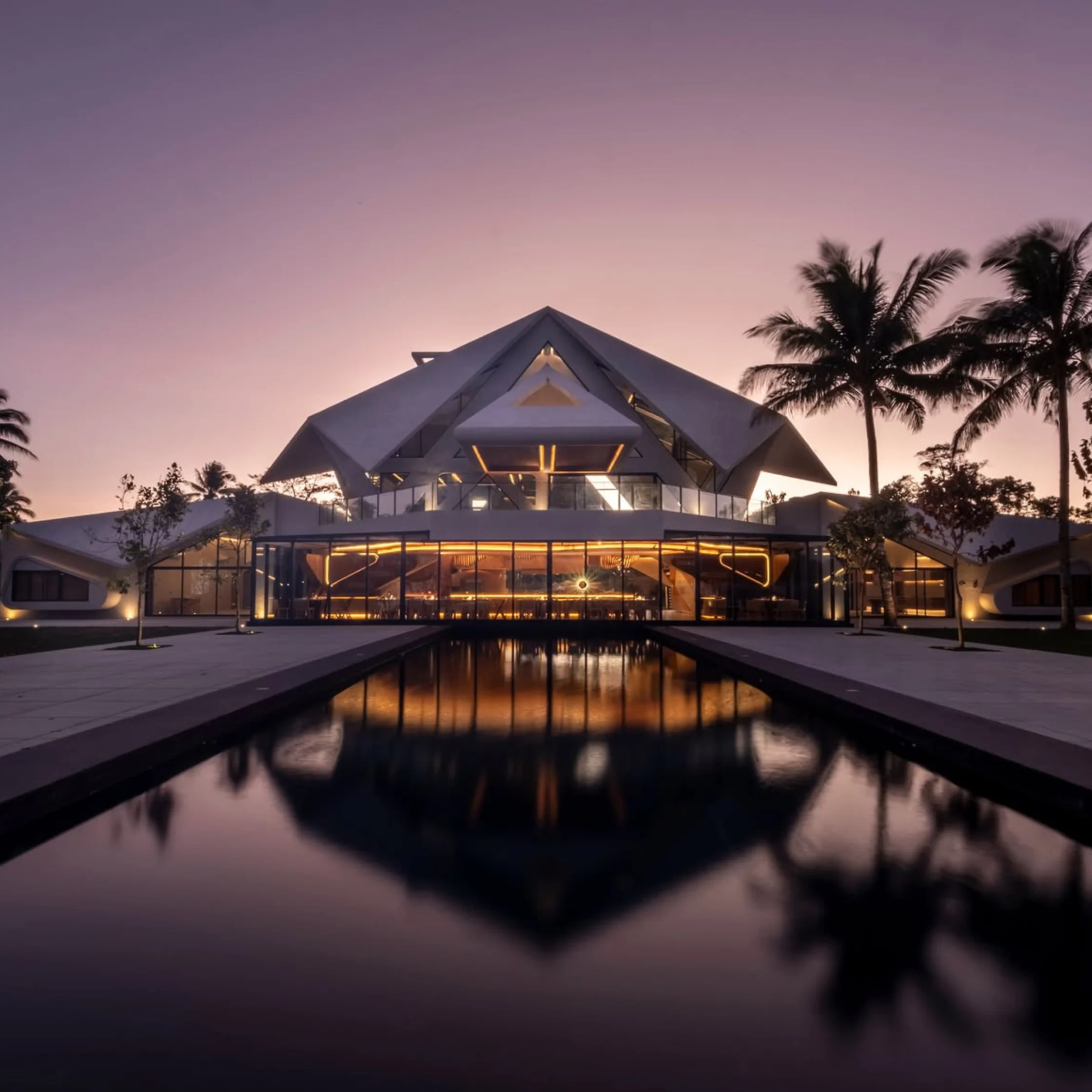 Modern glass building with geometric roof, warm interior lights, palm trees on the right, and a reflecting pool at sunset.