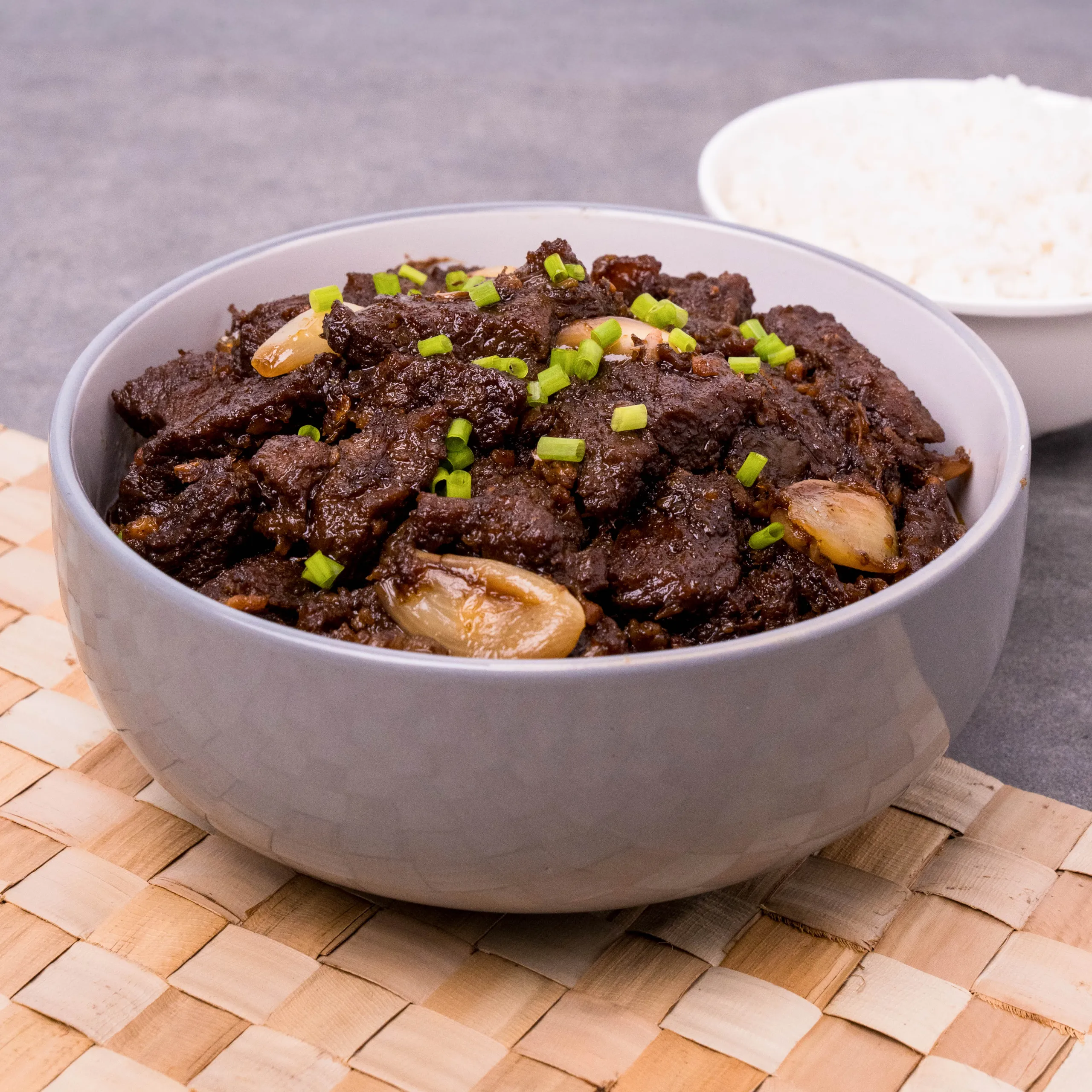 Bowl of dark braised beef with whole onions and chopped green onions on top, set on a wooden table with bread in the background