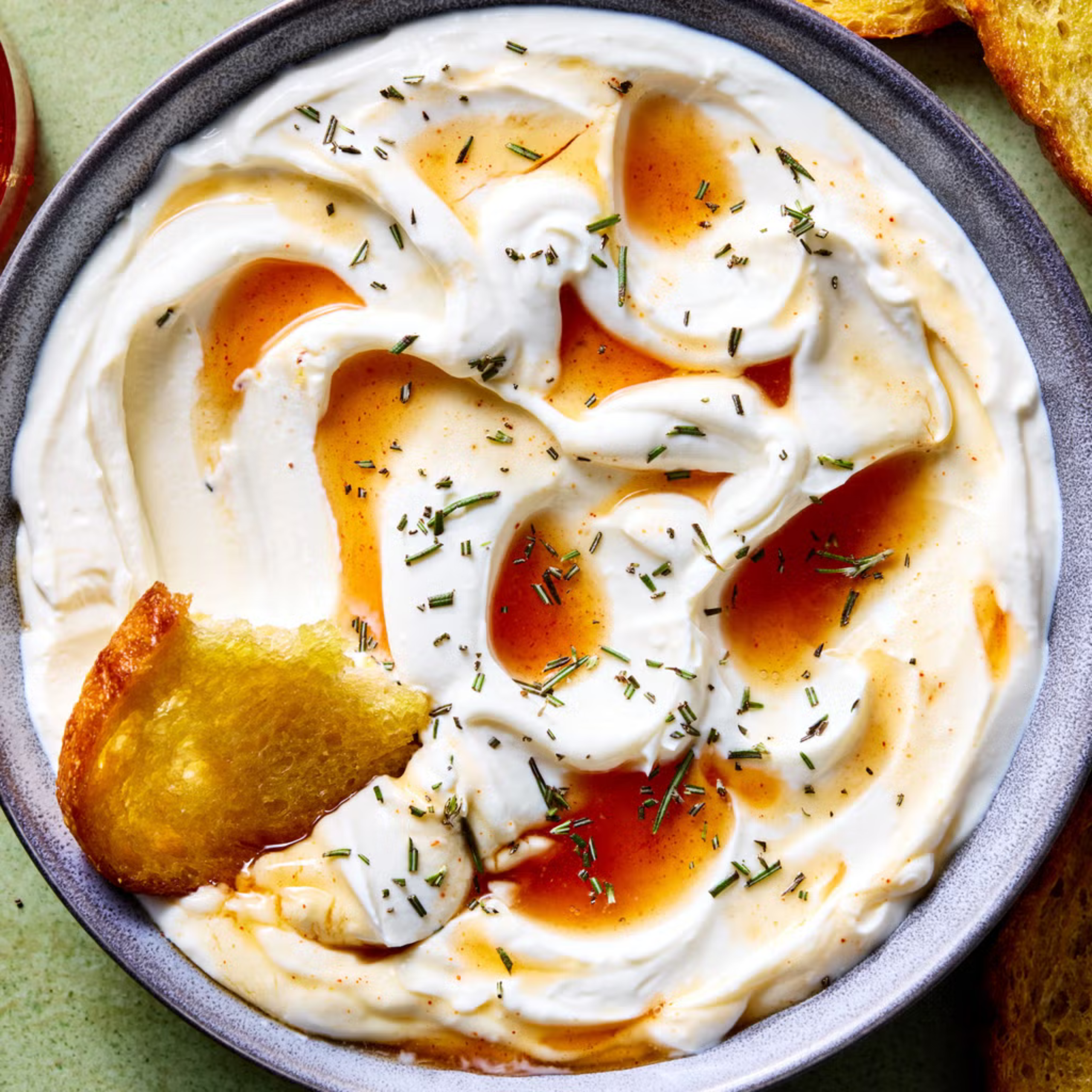 Close-up of a creamy white dip (yogurt/mascarpone) with olive oil drizzle and sprinkled chopped rosemary on a plate, plus a crisp bread piece.