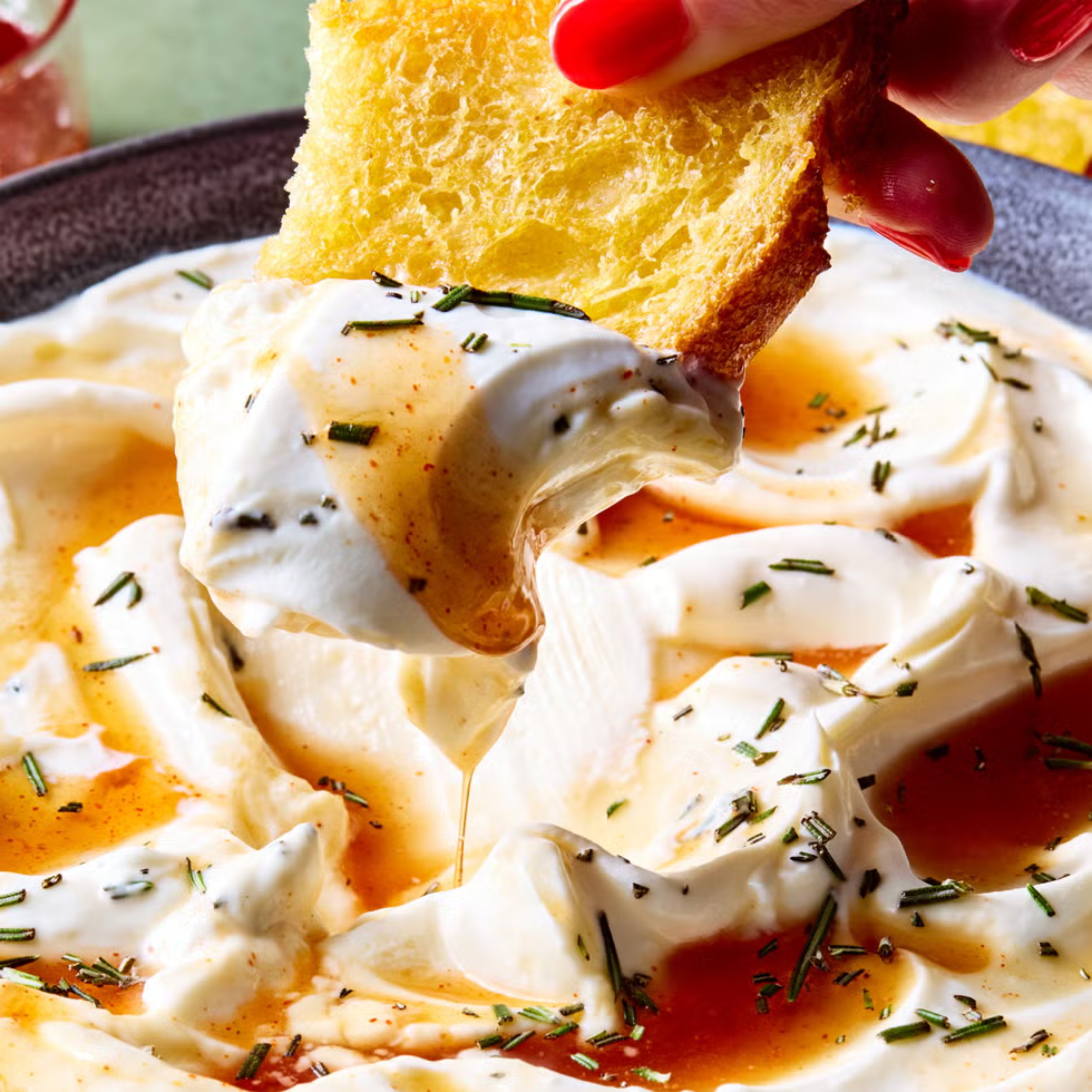 Close-up of a crusty bread slice being dipped into a creamy white herb sauce with chopped chives sprinkled on top.