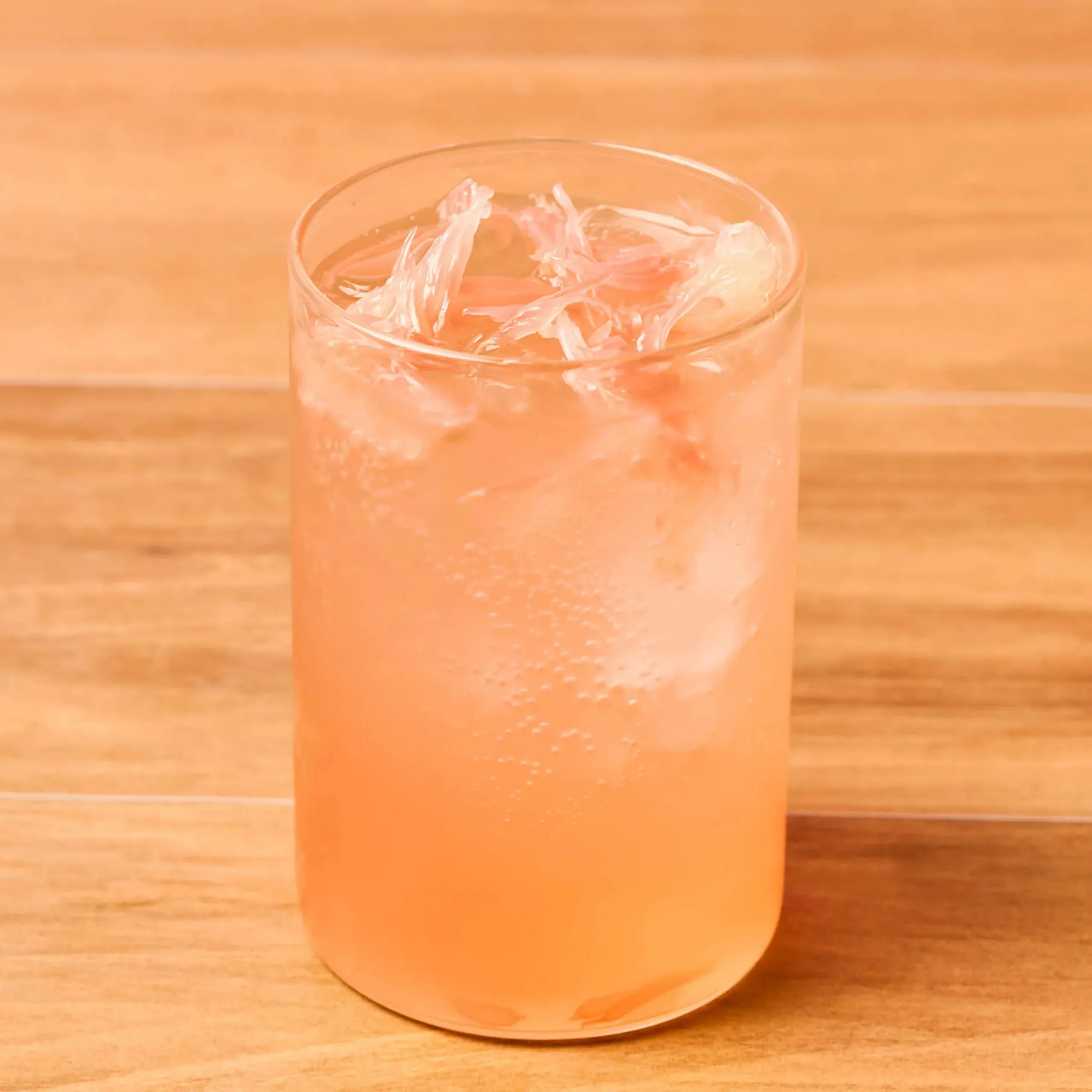 Close-up of a glass of orange-pink juice with ice on a wooden table, sunlight glow behind it.
