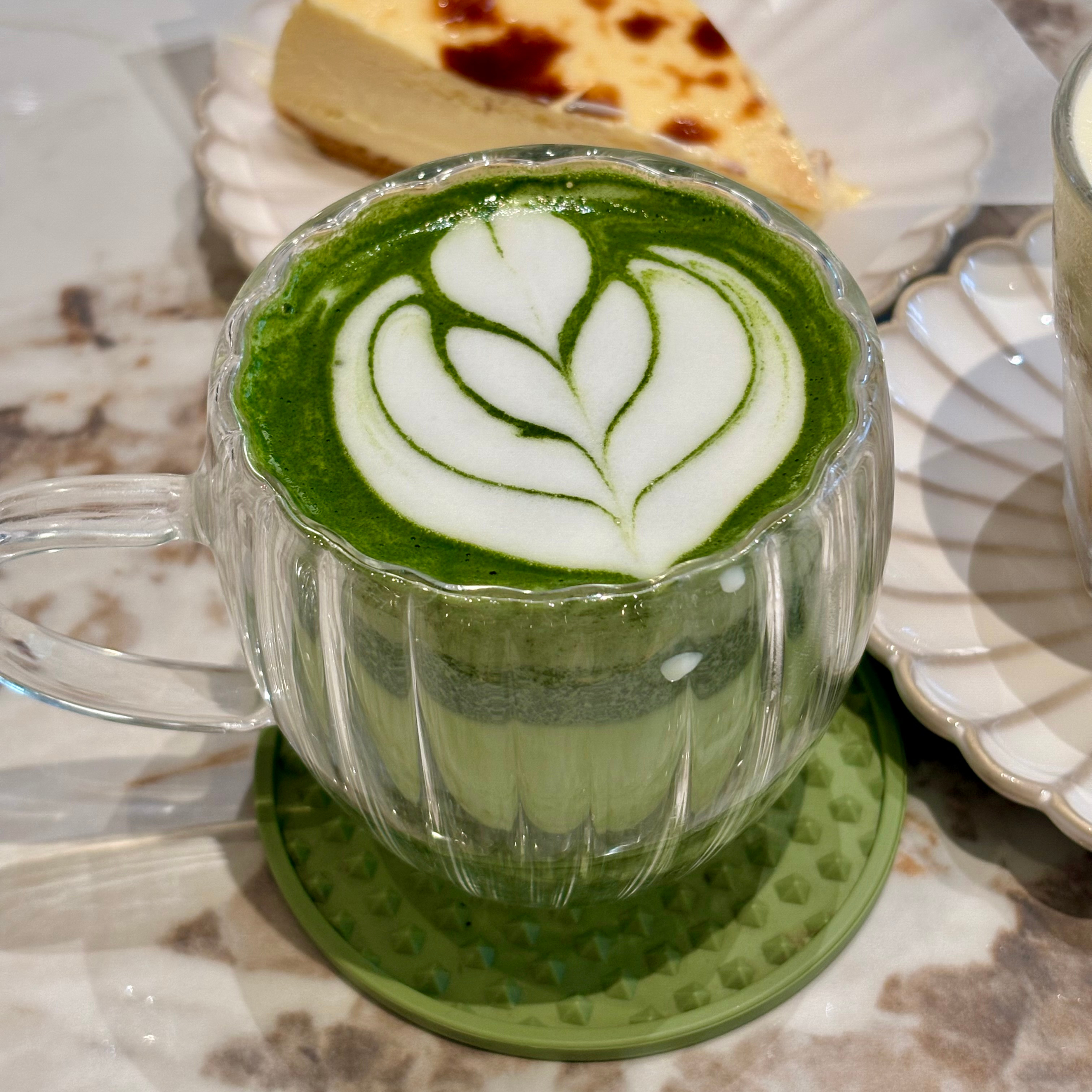 Green matcha latte with white heart latte art in a glass cup on a table, cake slice blurred in background