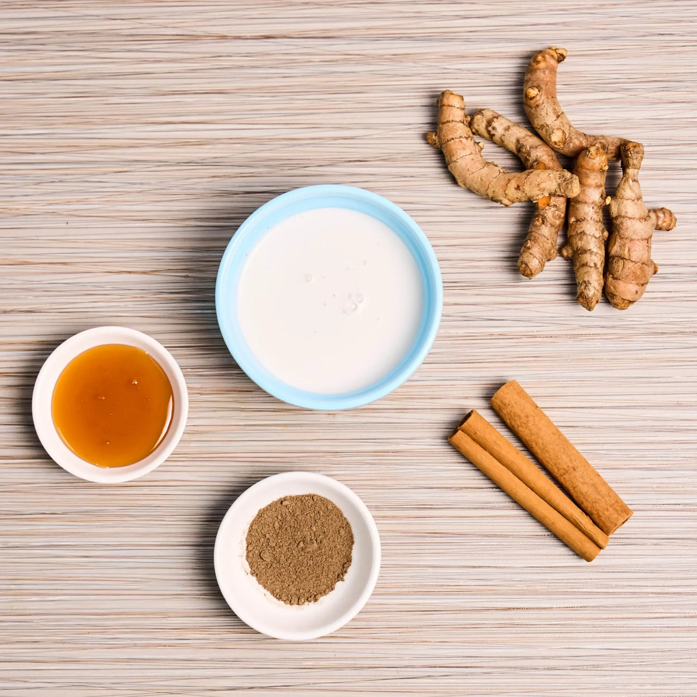 Turmeric roots and cinnamon sticks beside bowls of yogurt and honey on a light wooden table surface.