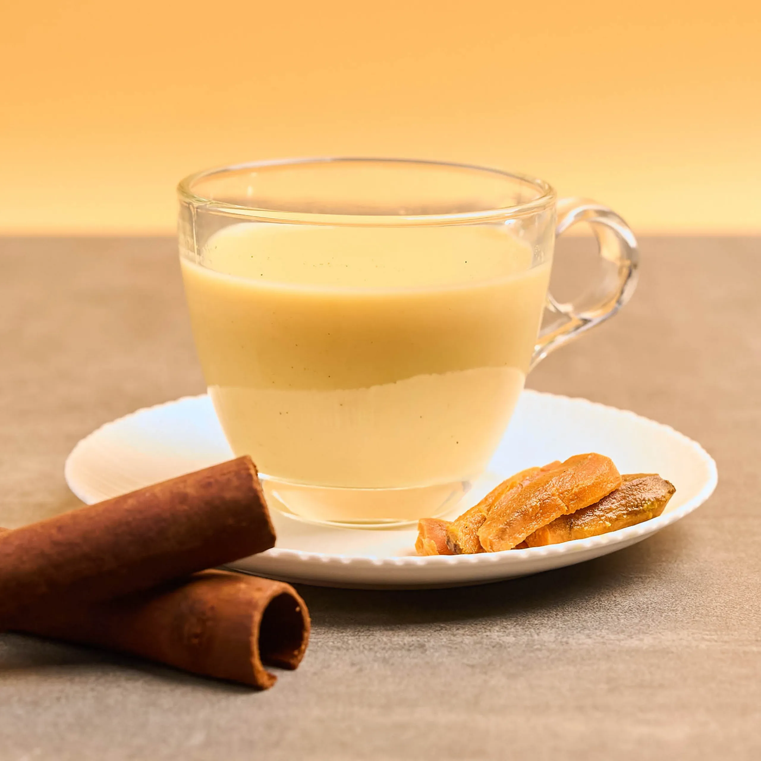 Close-up of a clear glass mug with a warm, light beverage, placed beside a plate with cinnamon sticks and biscotti.