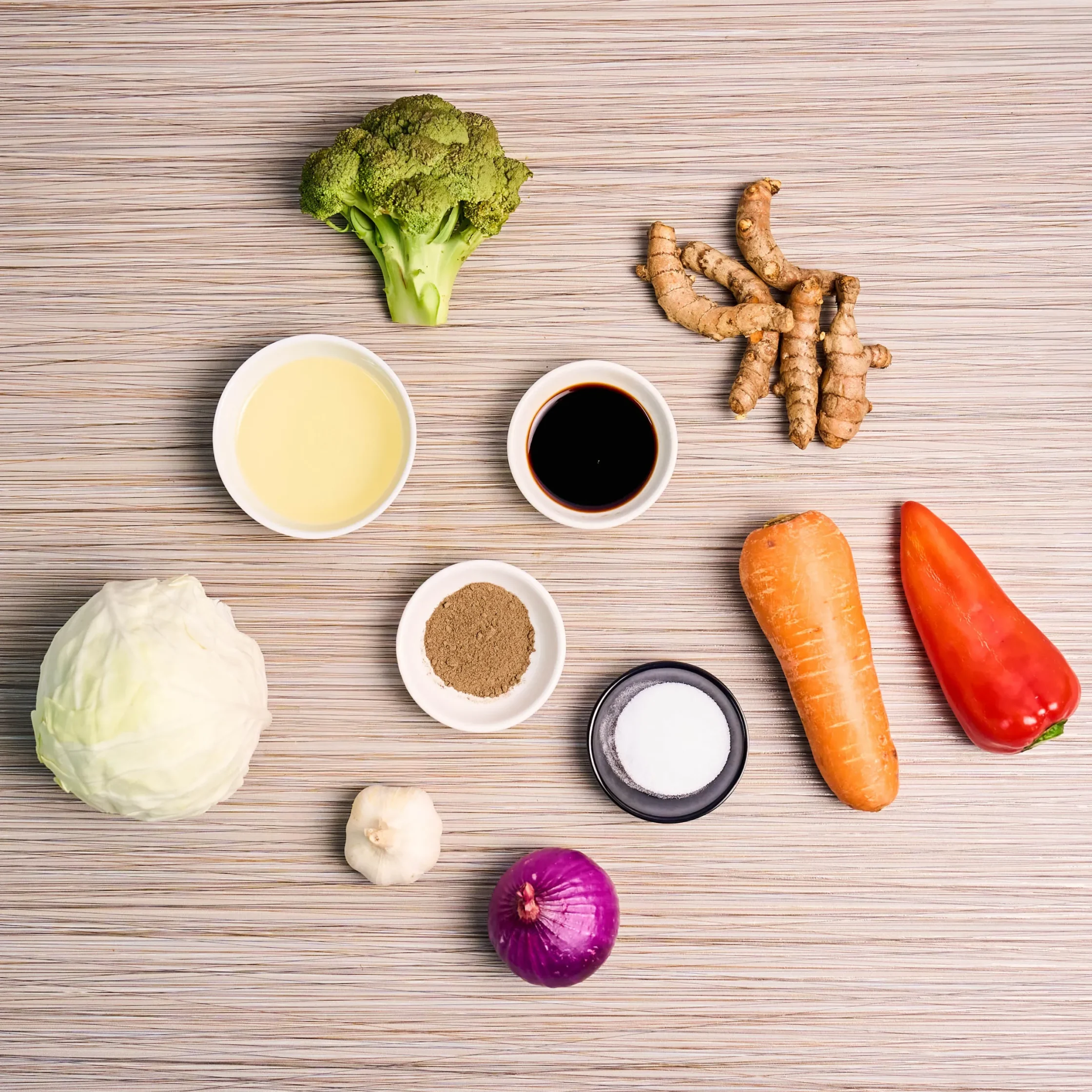 Top-down view of fresh vegetables and sauces on a wooden table: broccoli, carrots, cabbage, and ginger with small bowls of condiments nearby.