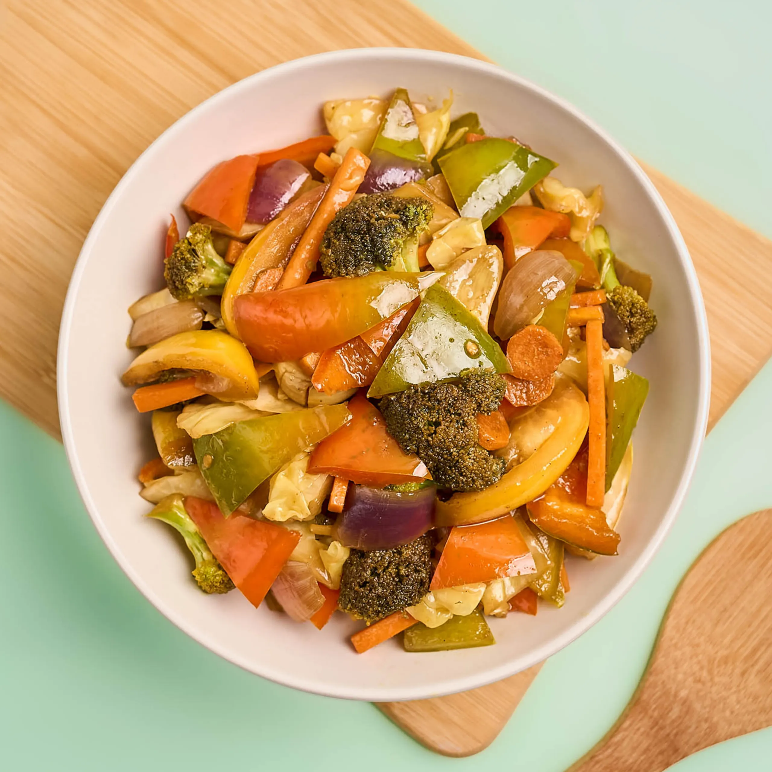 Assorted cooked vegetables in a white bowl, including broccoli, bell peppers, carrots, and onions, on a wooden surface.
