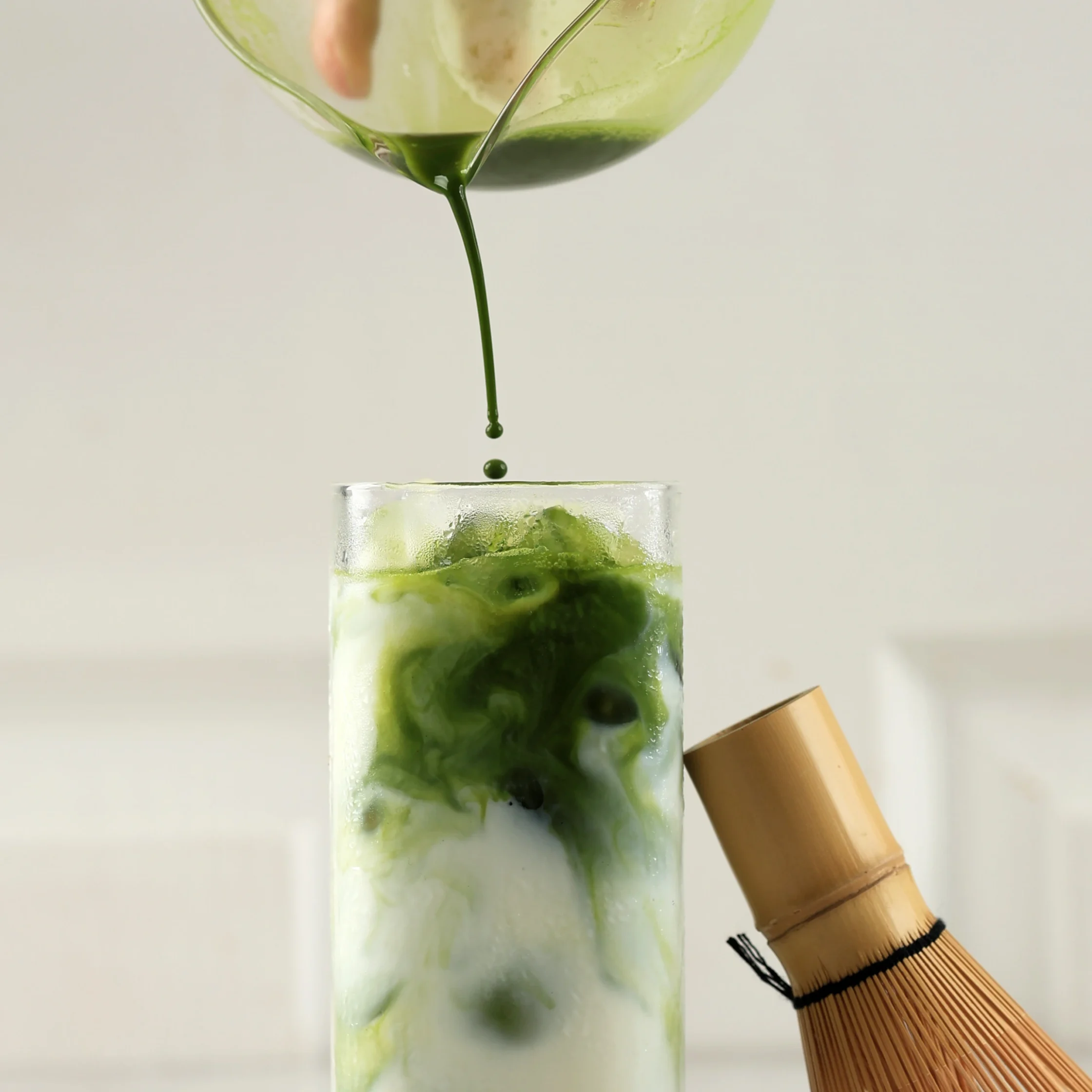 Green liquid matcha pours from a glass into a tall ice-filled glass as a bamboo tea whisk rests nearby.