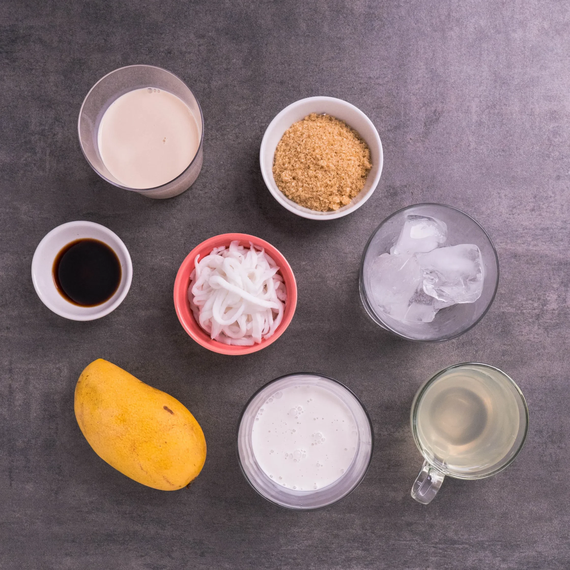 Top-down view of ingredients on a gray surface: glass of milk, bowl of brown sugar, bowl of sliced onions, small dish of soy sauce, yellow mango, ice in a clear pitcher, and a bowl of water.