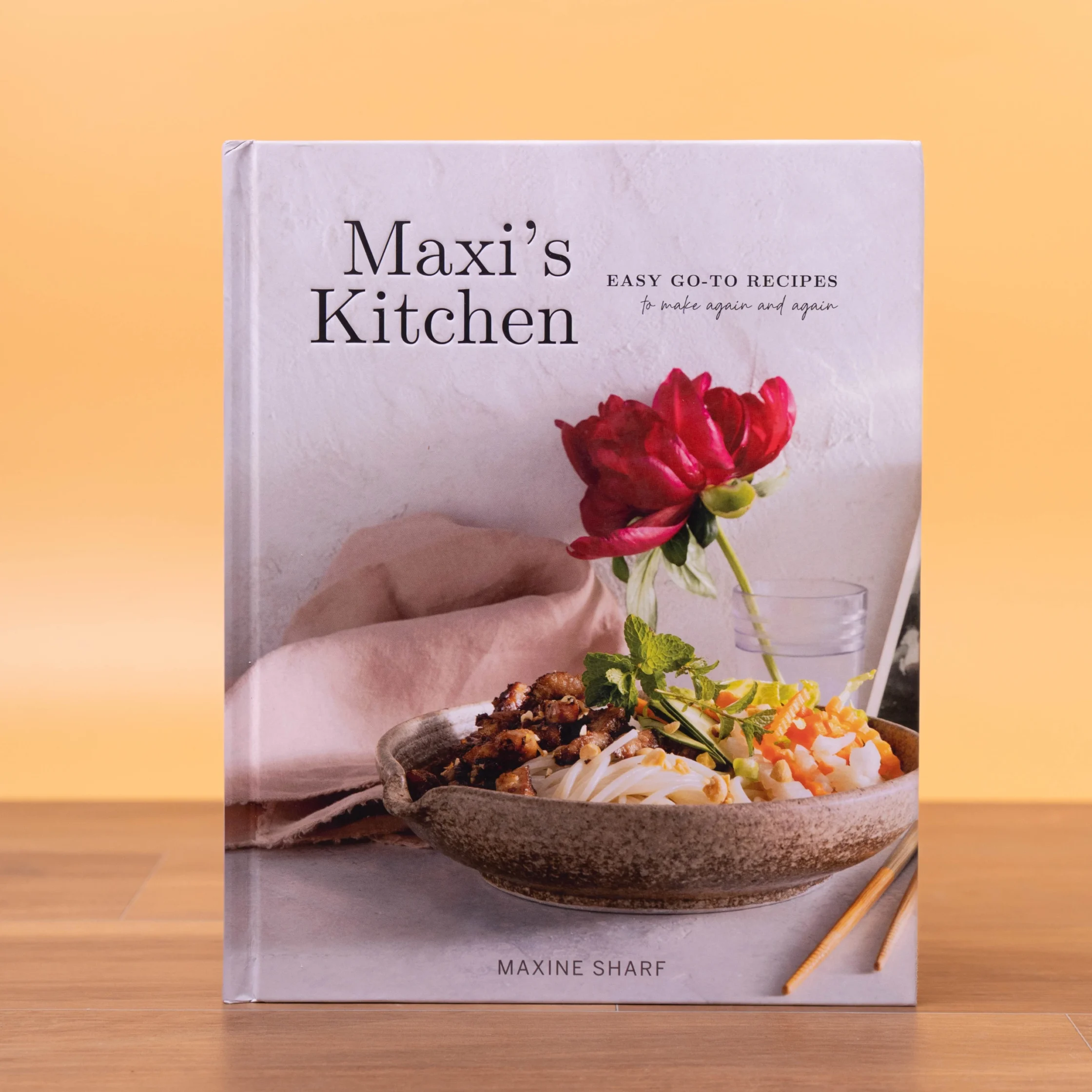 Cookbook cover showing the title Maxi's Kitchen with a red flower bouquet and a hand arranging food on a plate, chopsticks on the table behind.
