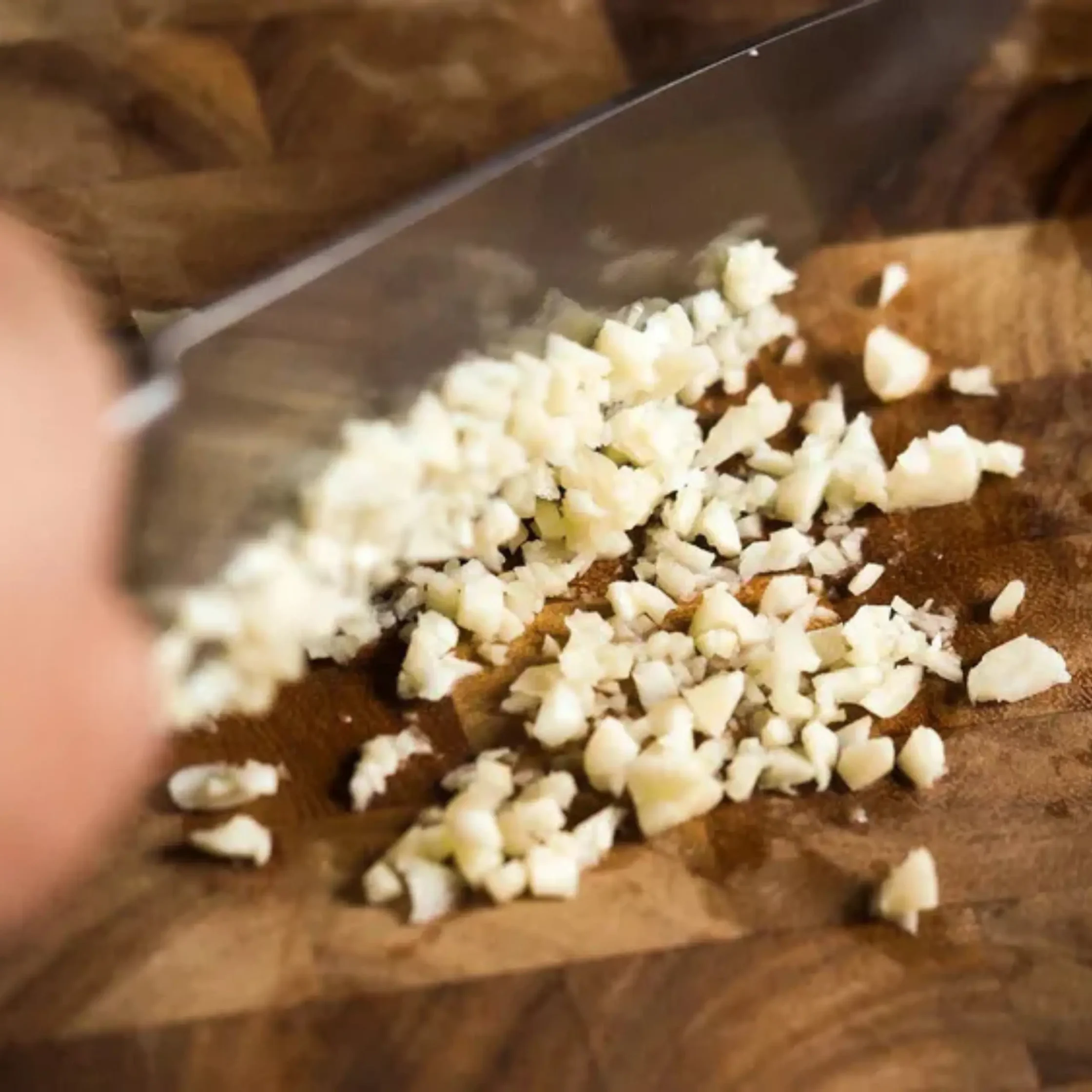 Chopped garlic scattered on a wooden cutting board beside a chef's knife tip visible
