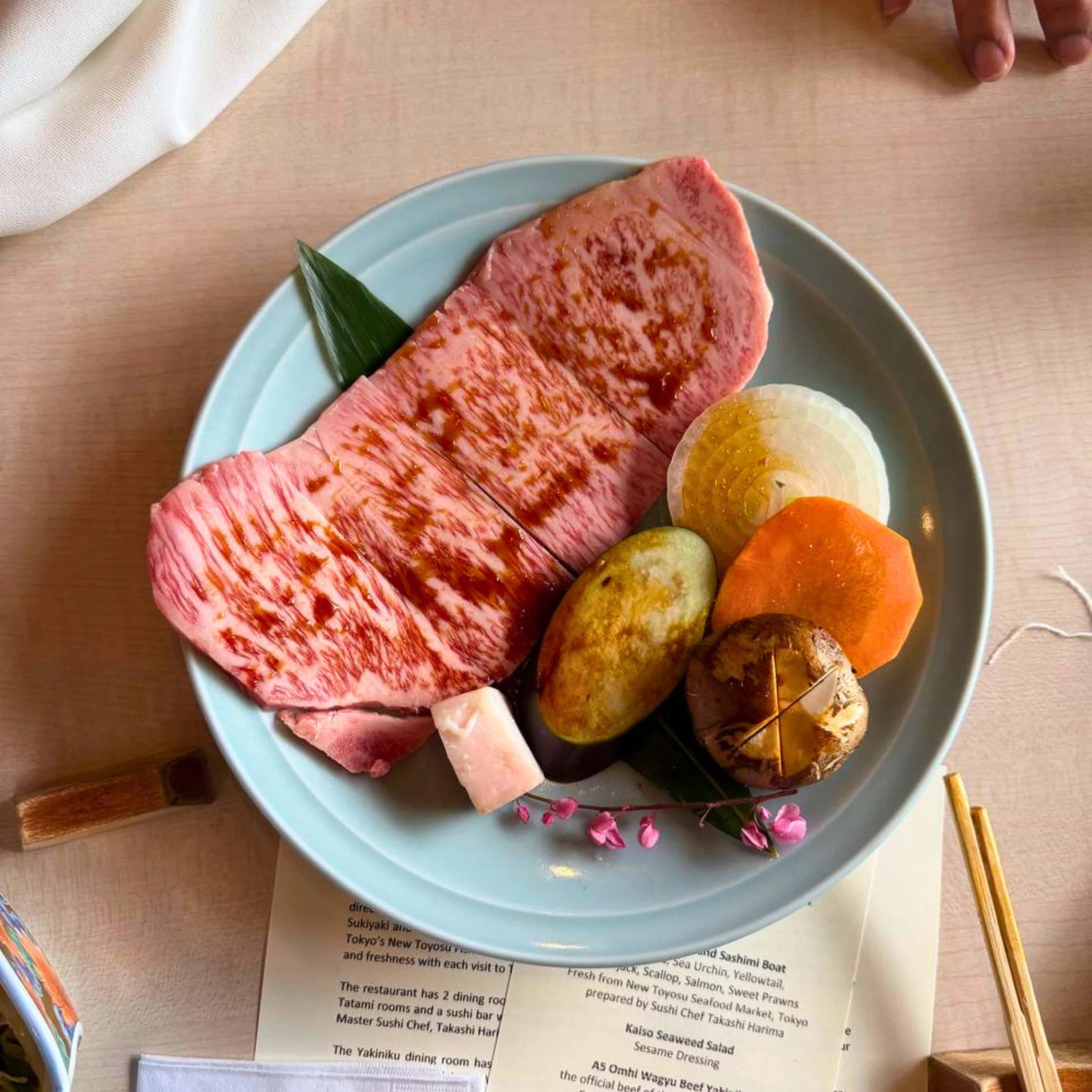 Thinly sliced marbled beef on a pale blue plate with a green leaf and onion slices on a wooden table, menu visible underneath omi beef rosu and vegetables ready for the grill