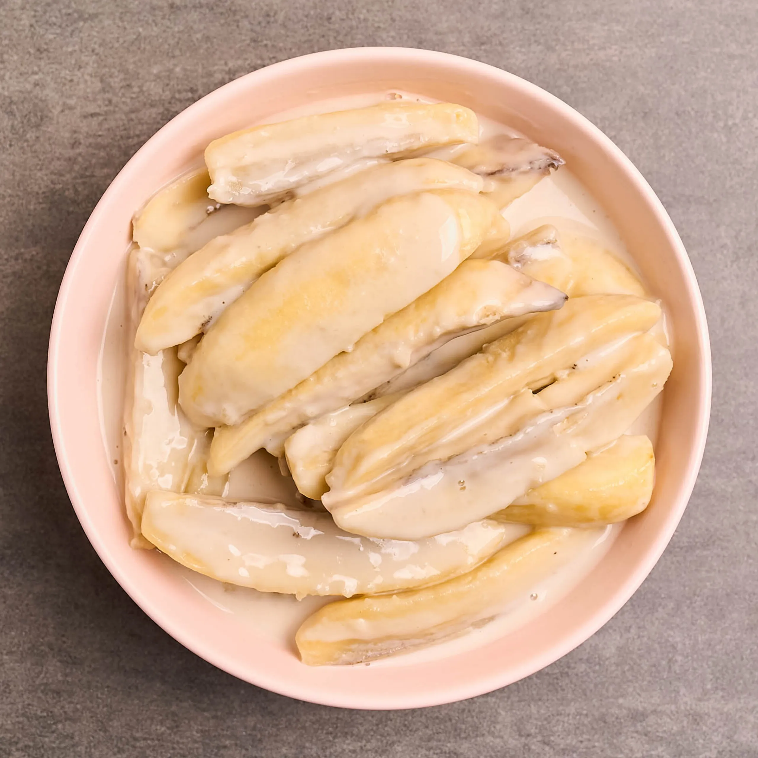 Sliced pale yellow bananas arranged in a pink bowl with a white sauce coating them, seen from above on a gray surface