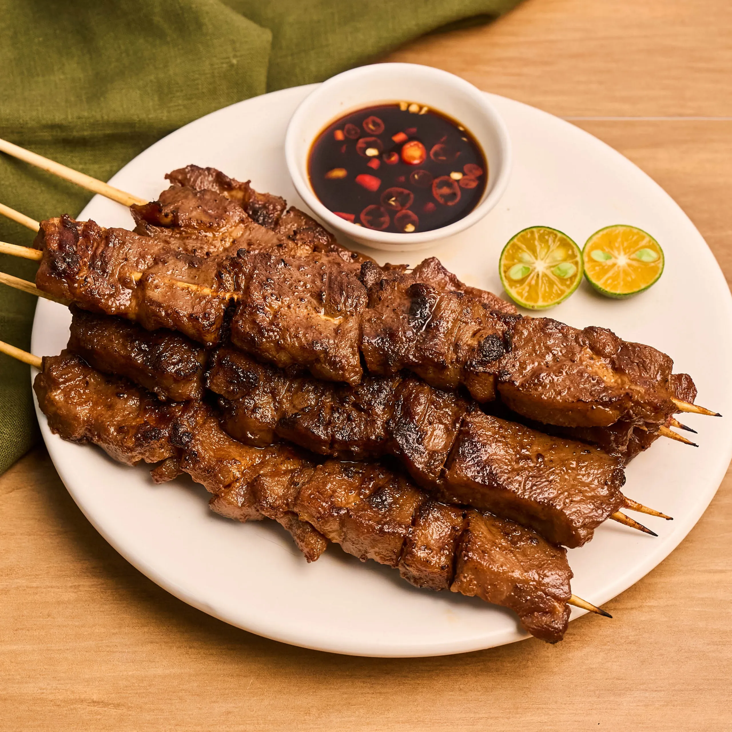 Skewered grilled beef with a dark dipping sauce in a small bowl and lime wedges on a white plate.