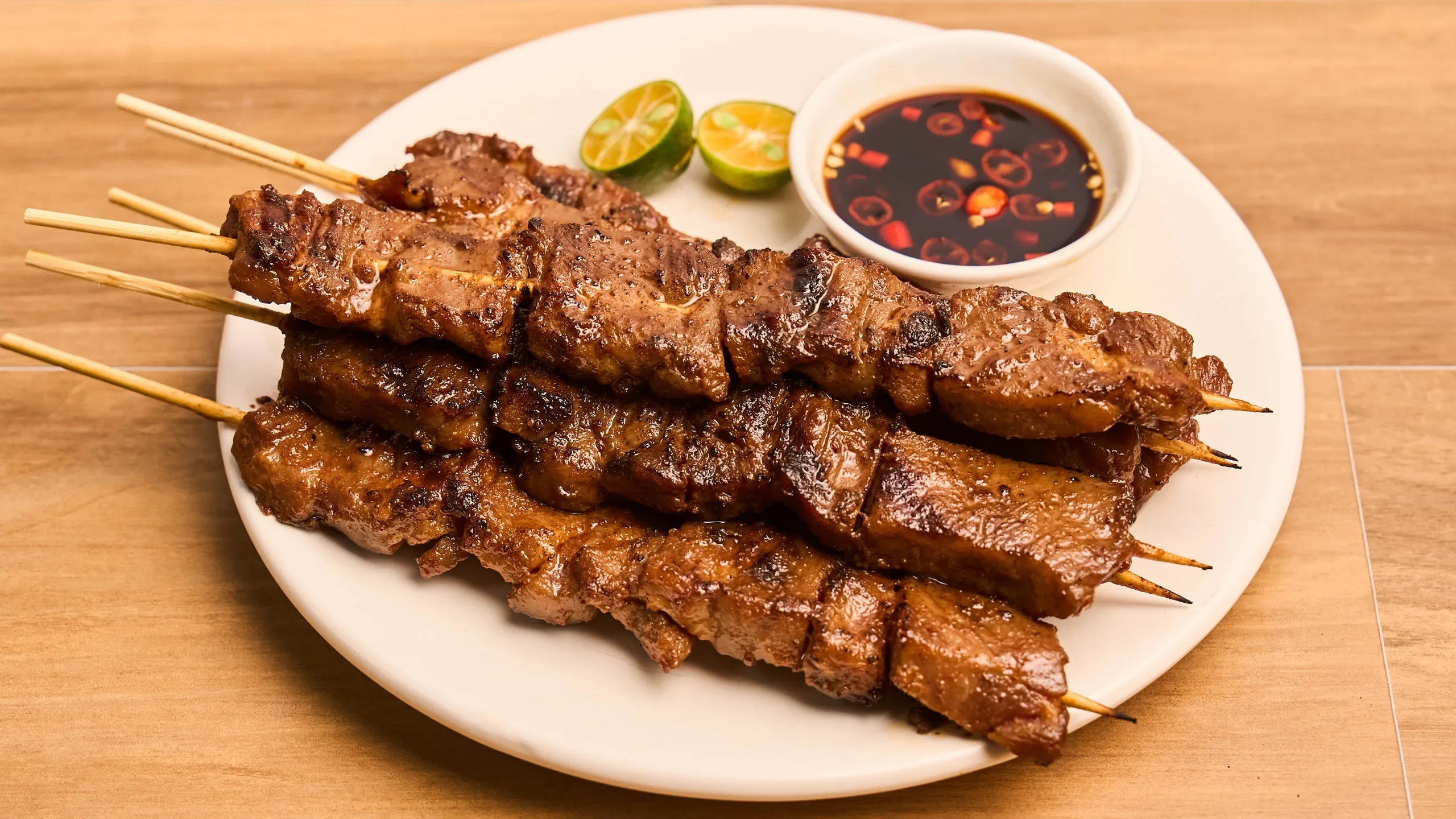 Skewered grilled meat on a white plate with a small bowl of dipping sauce and lime wedges on the side.