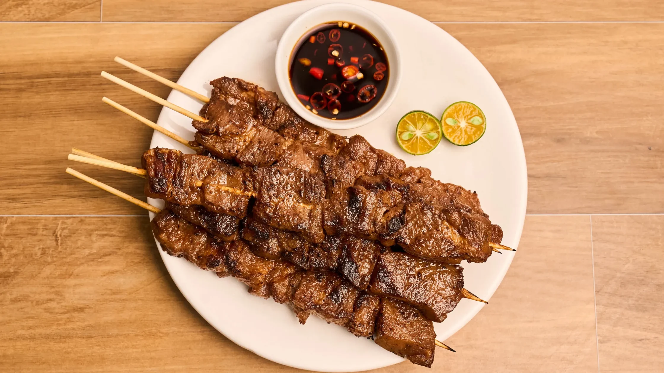 Pork skewers on a white plate with a dark dipping sauce in a small bowl and lime wedges on the side.