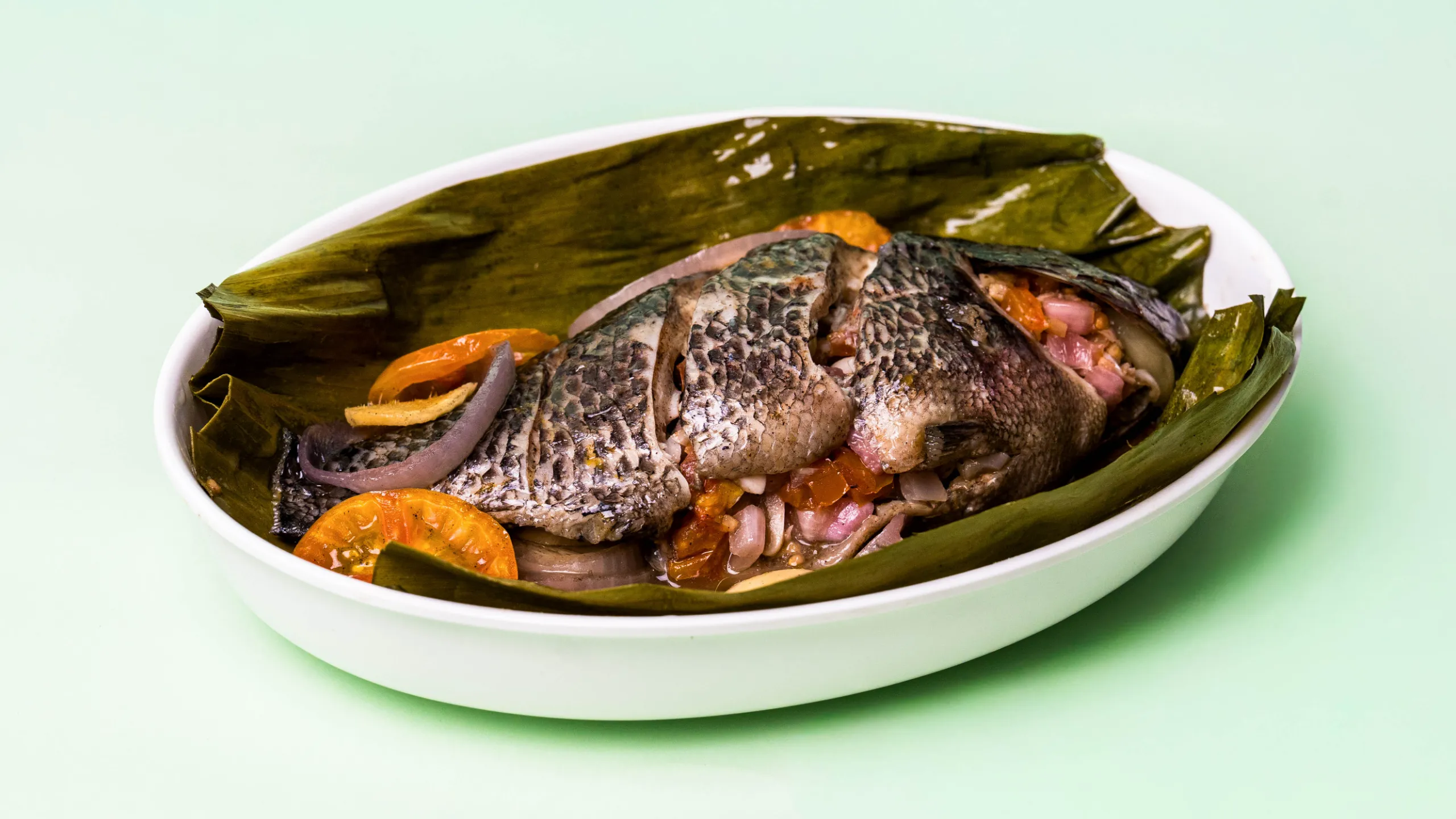 Whole grilled fish served in a banana leaf boat with onions and tomatoes on a white dish.