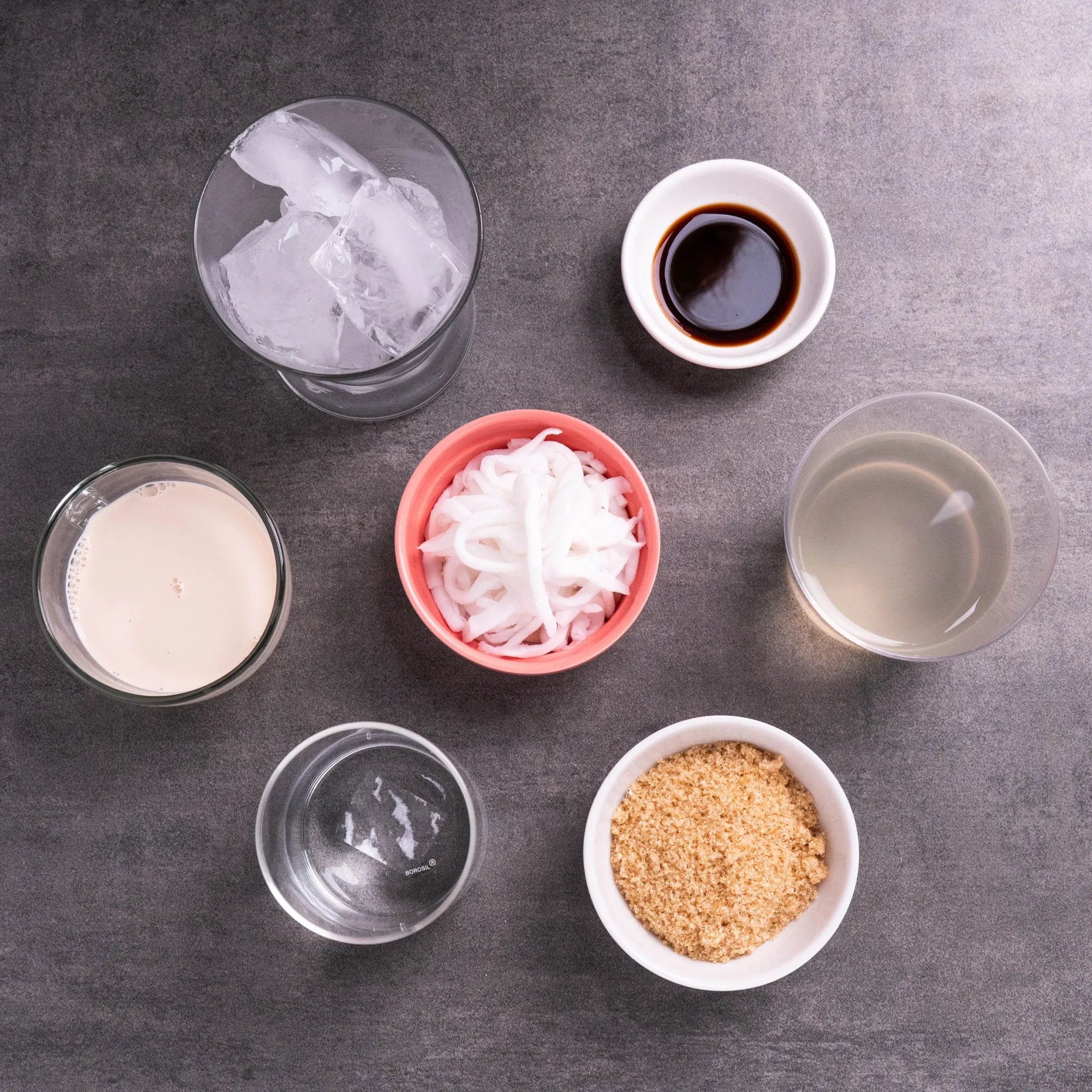 Top-down view of an ice-filled glass, a small cup of black coffee, and bowls of brown sugar and shredded coconut on a gray countertop.