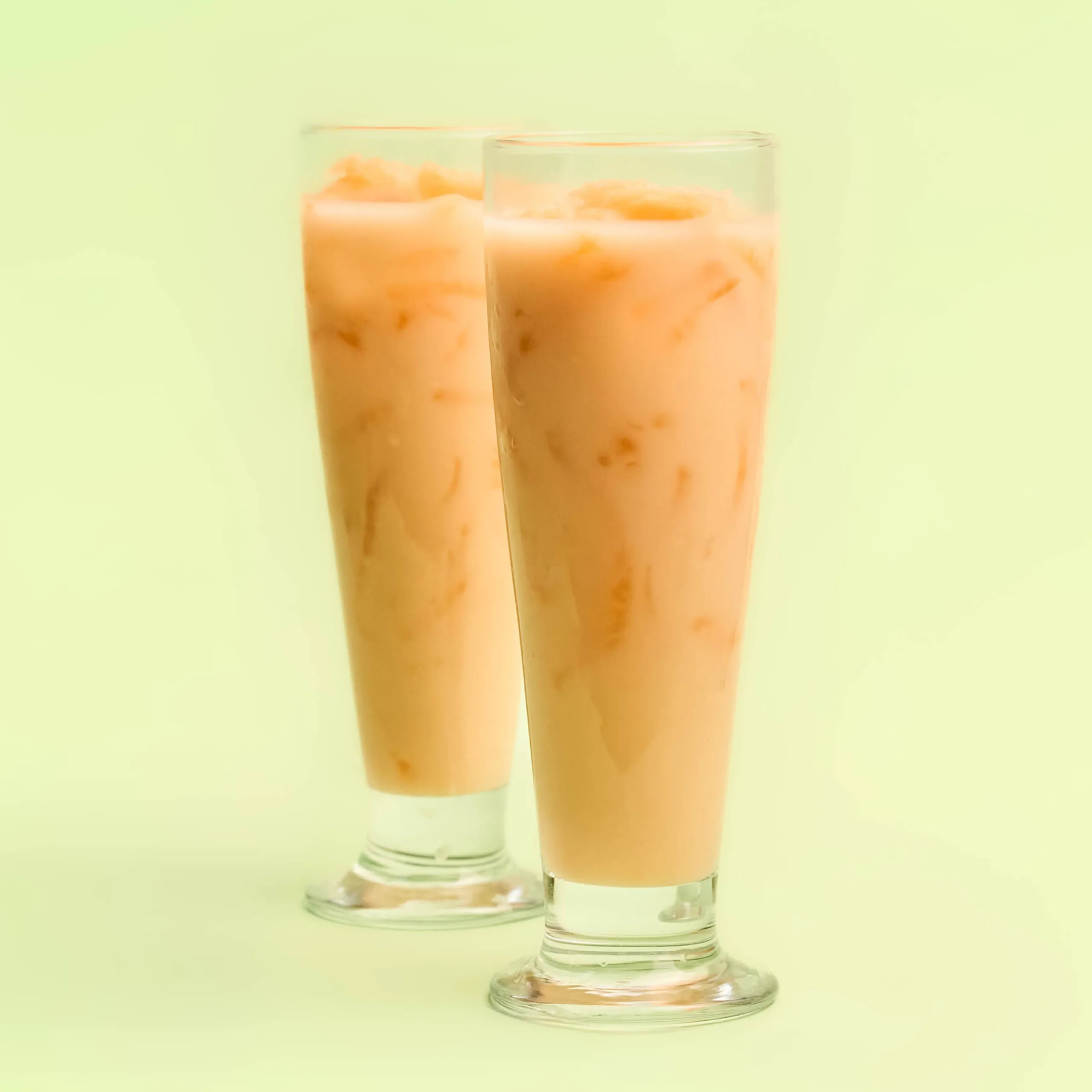 Two tall glasses of iced peach tea with ice against a pale yellow background.