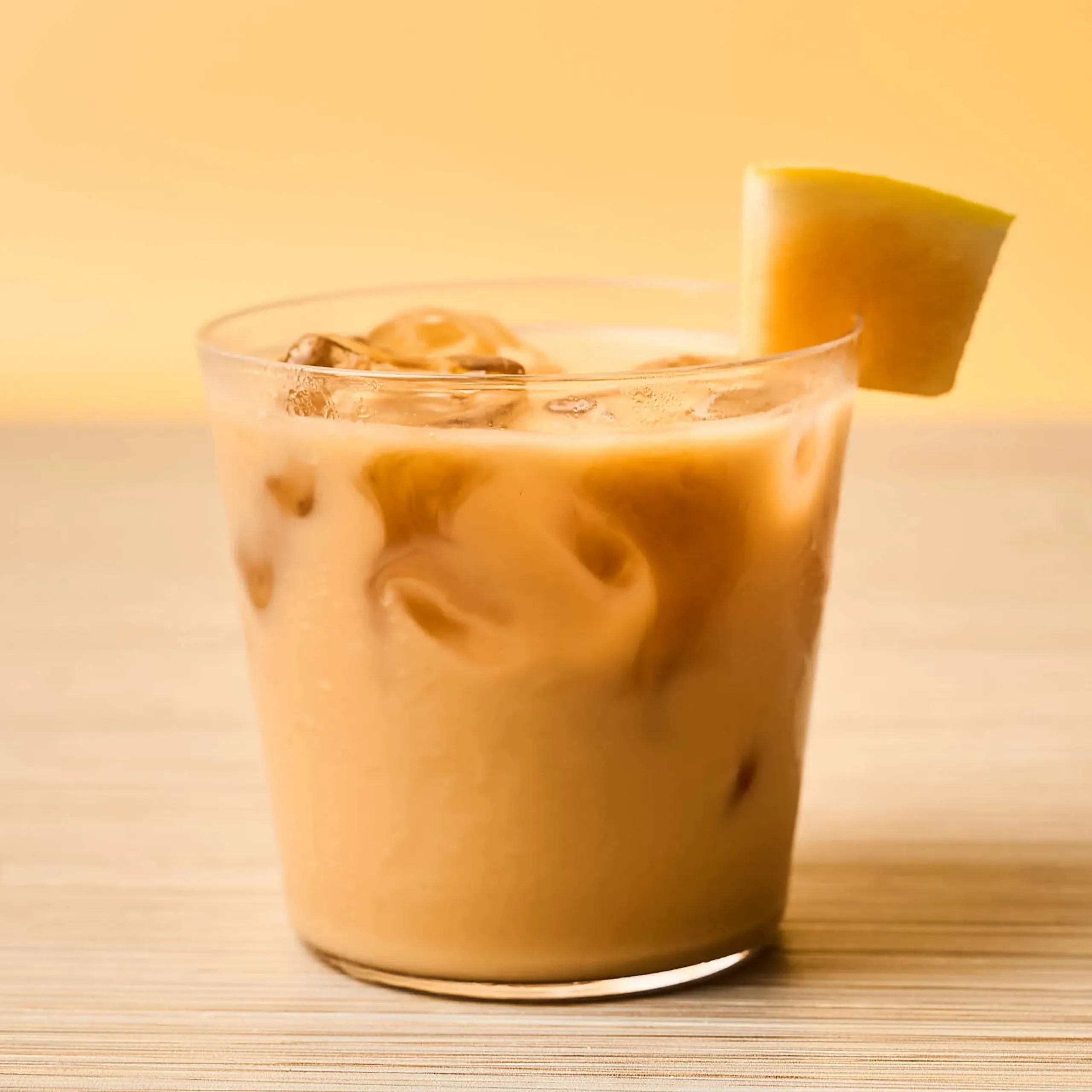 Glass of iced coffee with ice cubes and a lemon wedge on the rim, on a wooden surface.