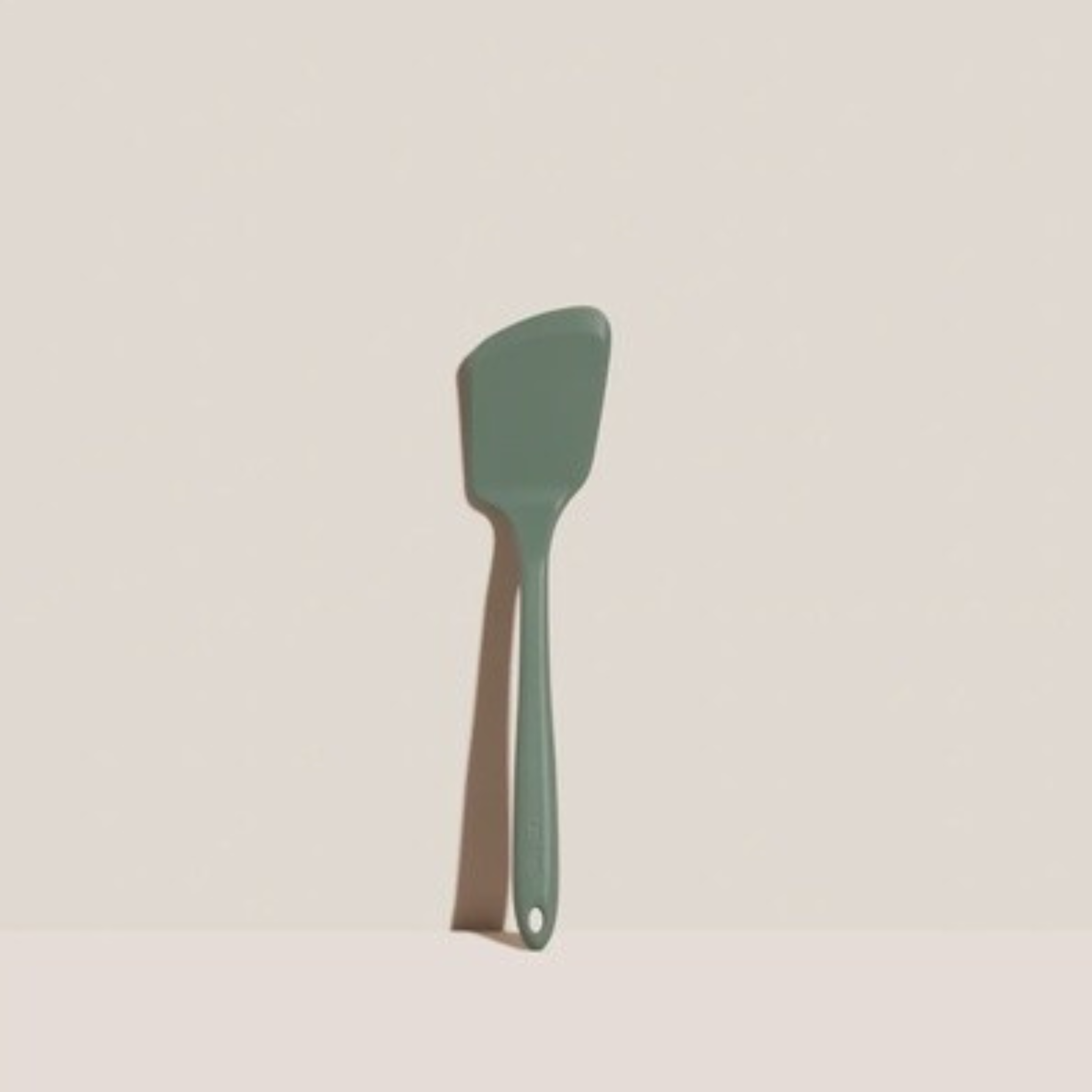 Green silicone spatula with a wooden handle hanging from a hook on a light beige wall.