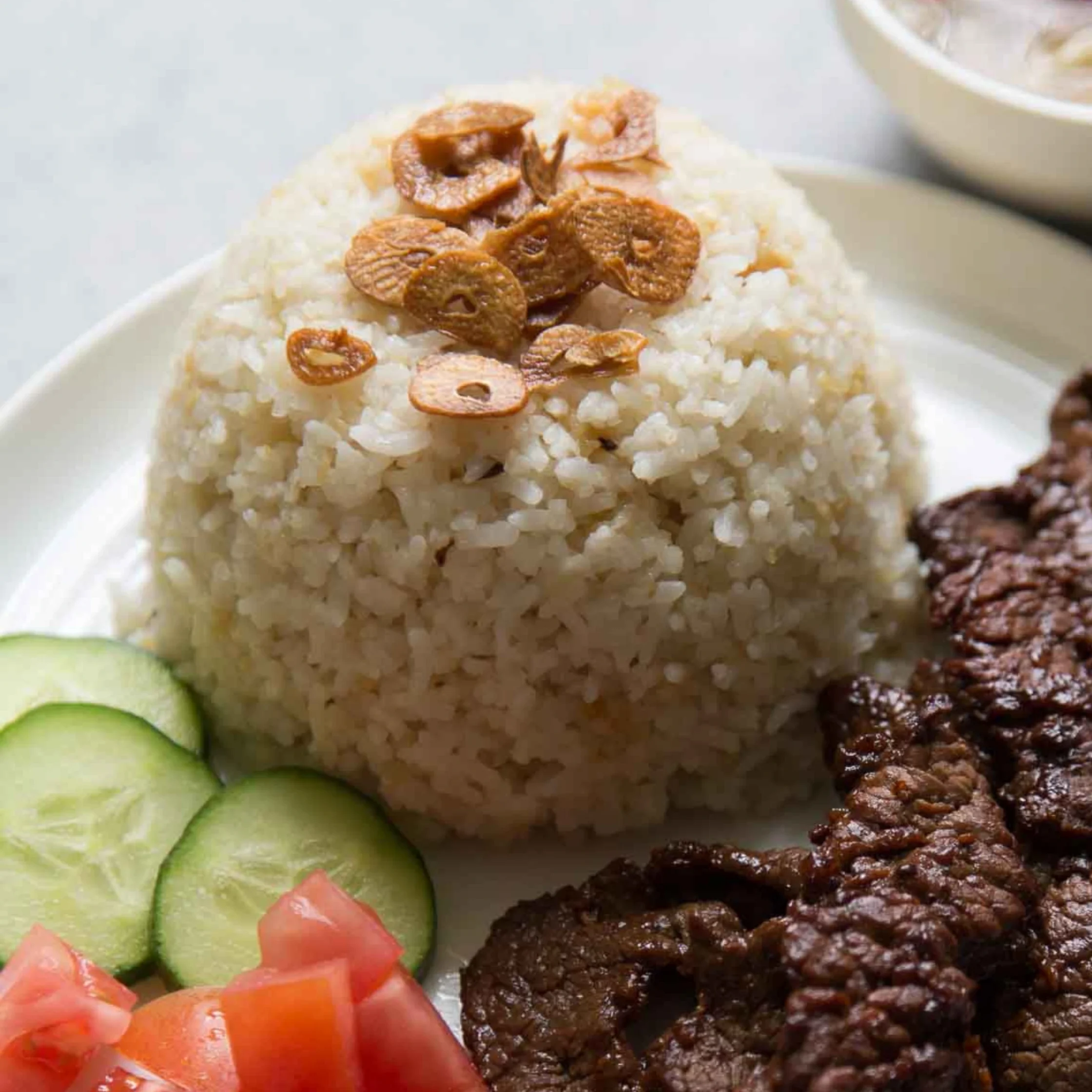 Close-up of a mound of white rice topped with fried shallots, with cucumber slices and a beef patty on the side.