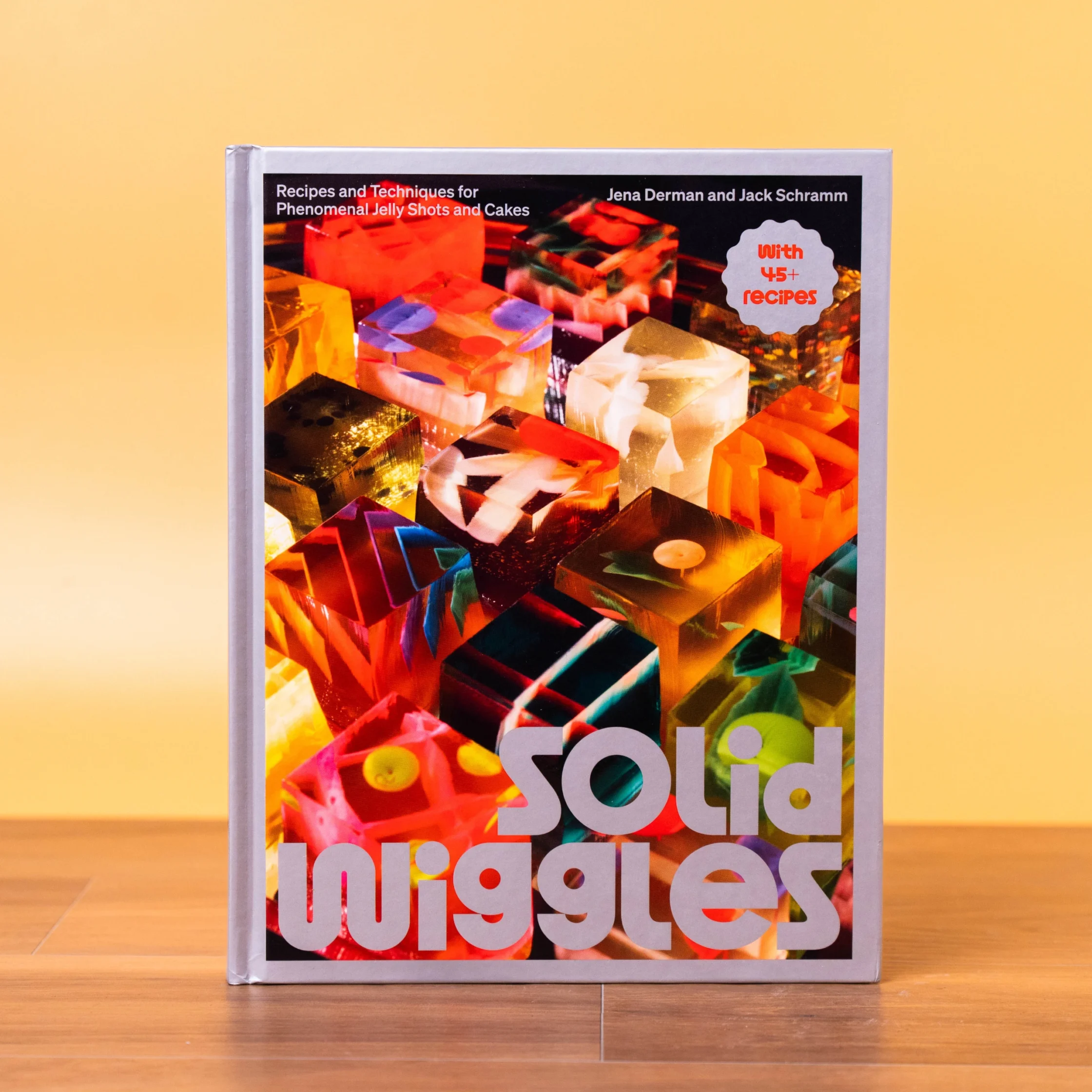 Colorful cookbook cover on a silver stand against a yellow background, titled 'Recipes and Techniques for Phenomenal Jelly Shots and Cakes' with authors Jena Derman and Jack Schramm.