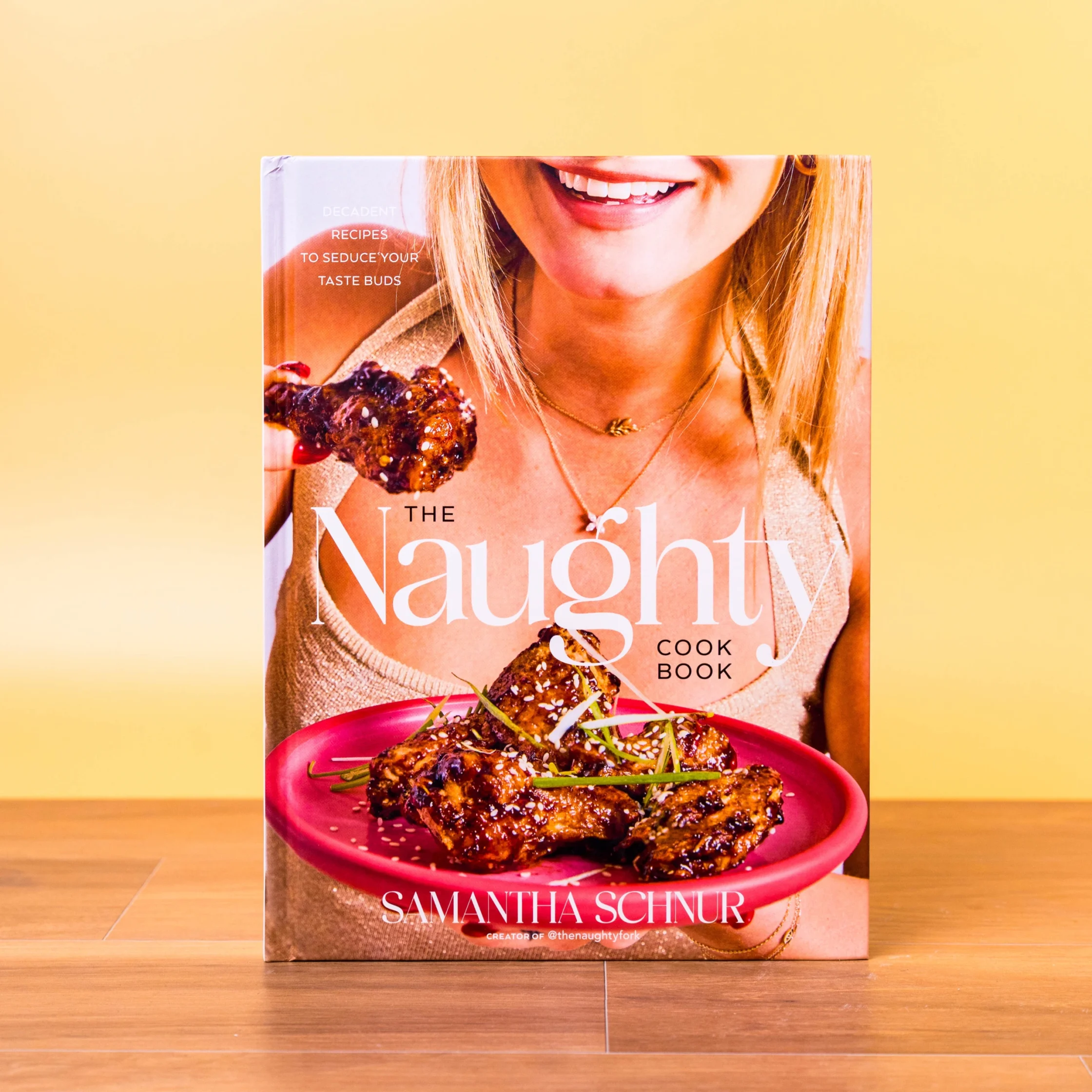 Close-up of a magazine cover on a wooden floor: a smiling woman holding a glazed chicken dish on a pink plate, with the title 'NAUGHTY' and 'SAMANTHA SCHNUR' visible.