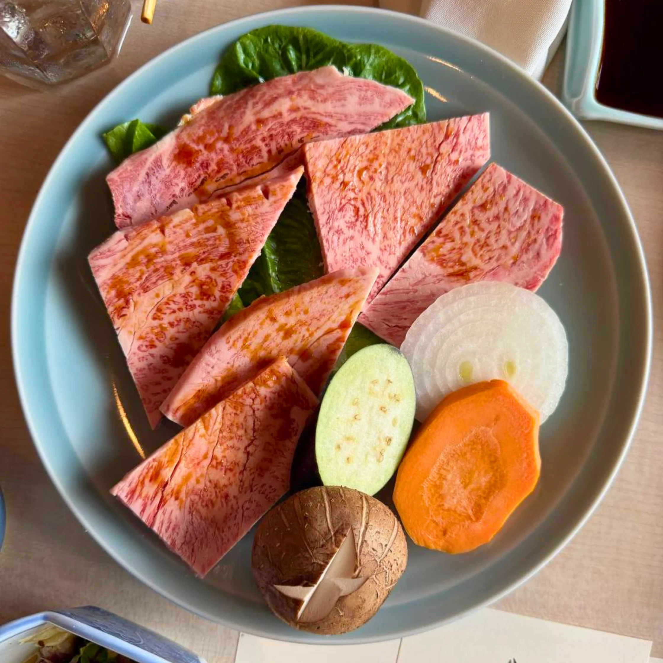Thinly sliced marbled beef arranged on a bed of greens on a blue plate with citrusy orange slices nearby. (Note: If used as decorative food platter, communicates a sliced beef dish the omi wagyu ribeye has more marbling one can ask for