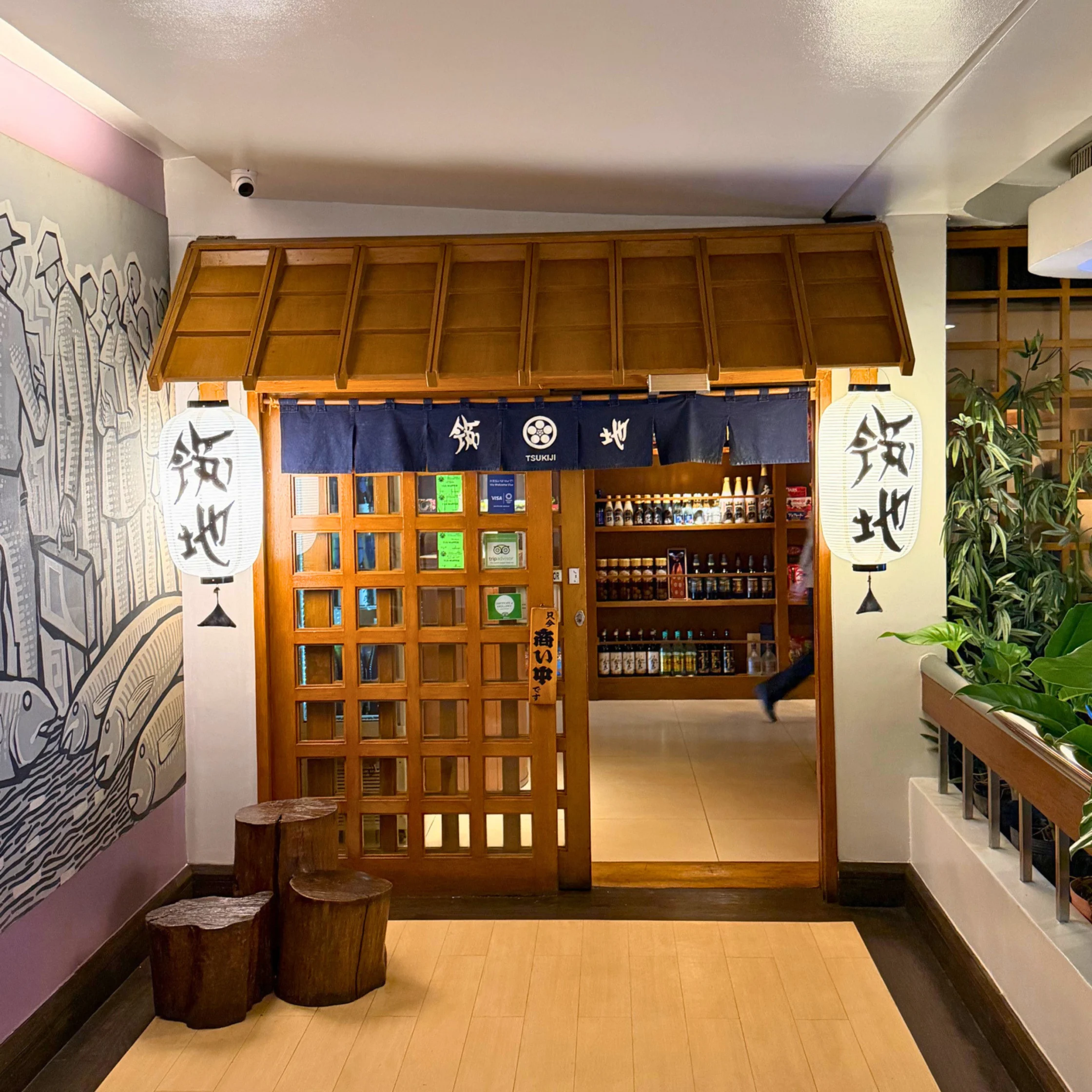 Entrance to a Japanese restaurant with wooden lattice doors, blue fabric curtain, and paper lanterns, plus a mural on the left wall.