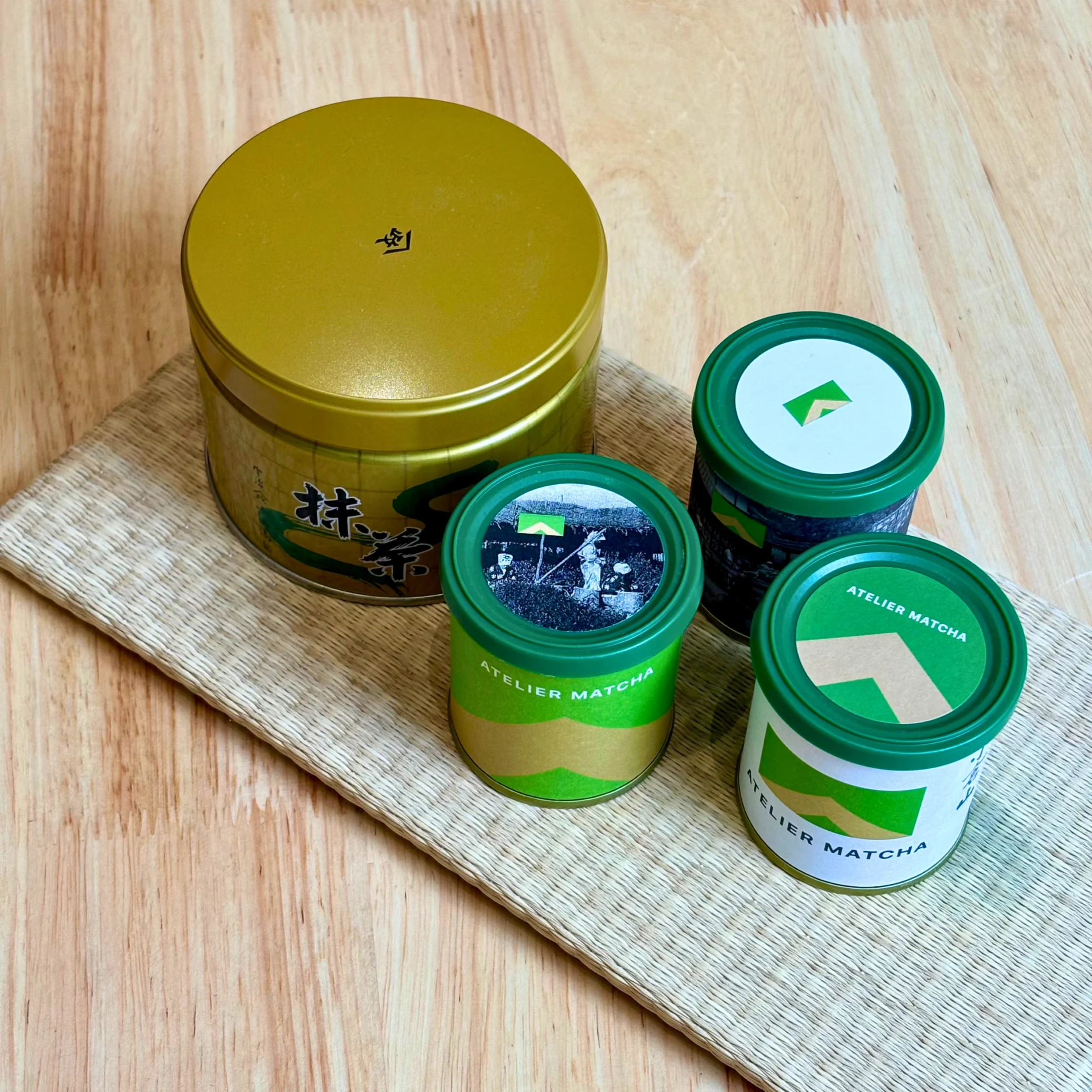 three small green tubs and one big golden tub of matcha placed on a light wooden table