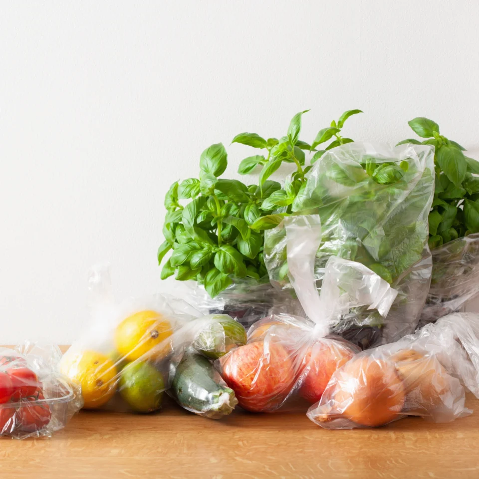 Assorted fruit in plastic bags on a wooden table, including apples, oranges, pears, and lemons, with a basil plant in the background.