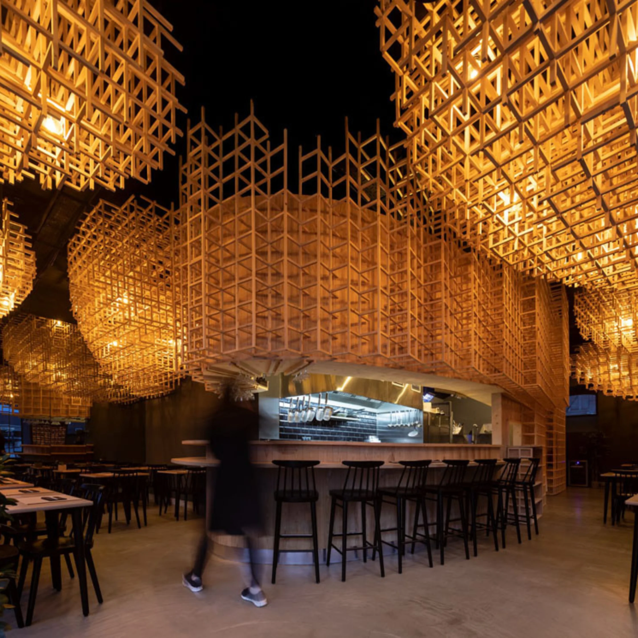 Interior dining area with geometric wooden lattice panels overhead, warm amber lighting, and a bar with stools in the foreground.