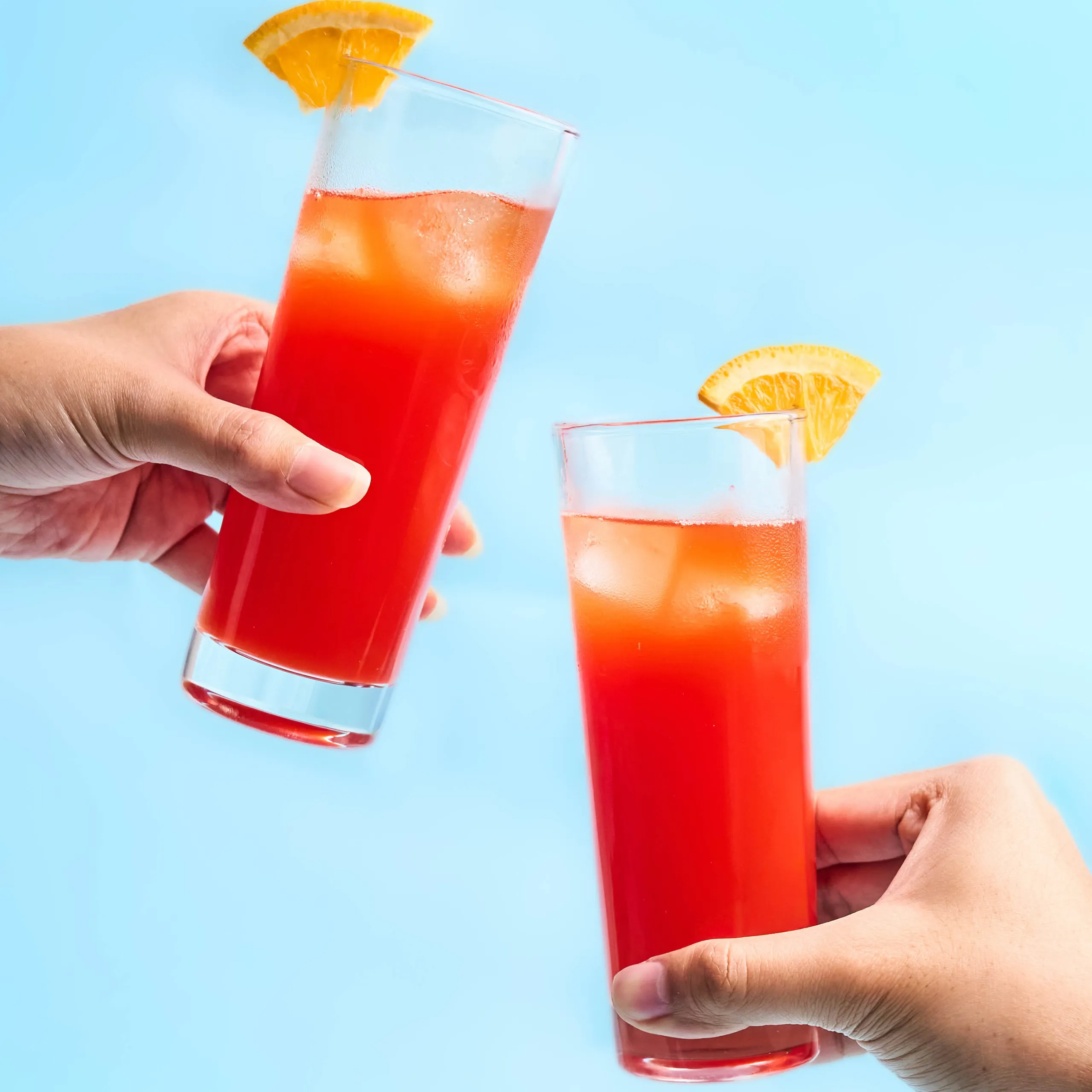 Hand holding a tall red cocktail in a glass with an orange wedge garnish against a blue background.