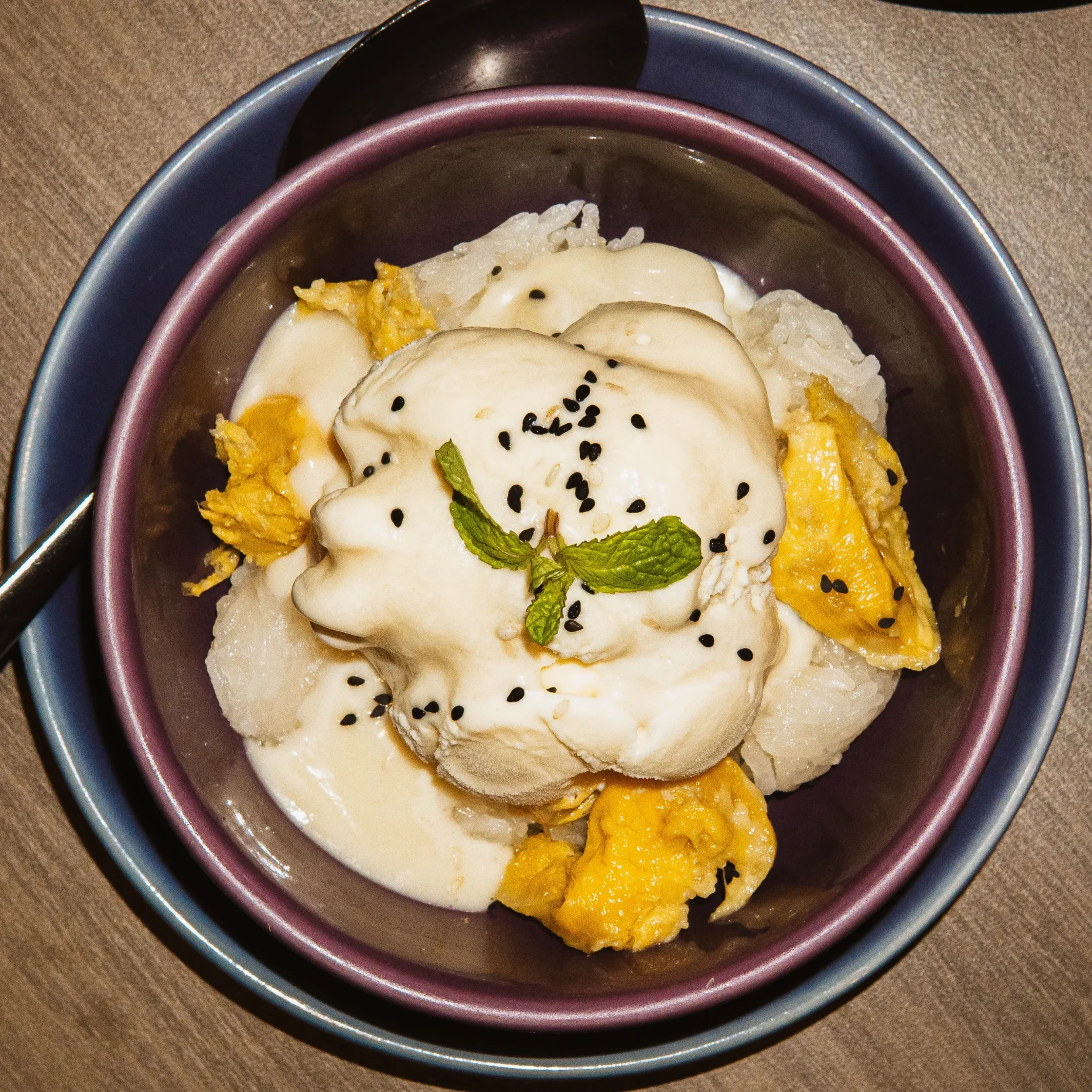 Close-up of a dessert with white creamy topping, black sesame seeds, mint leaf, and mango slices in stacked bowls.