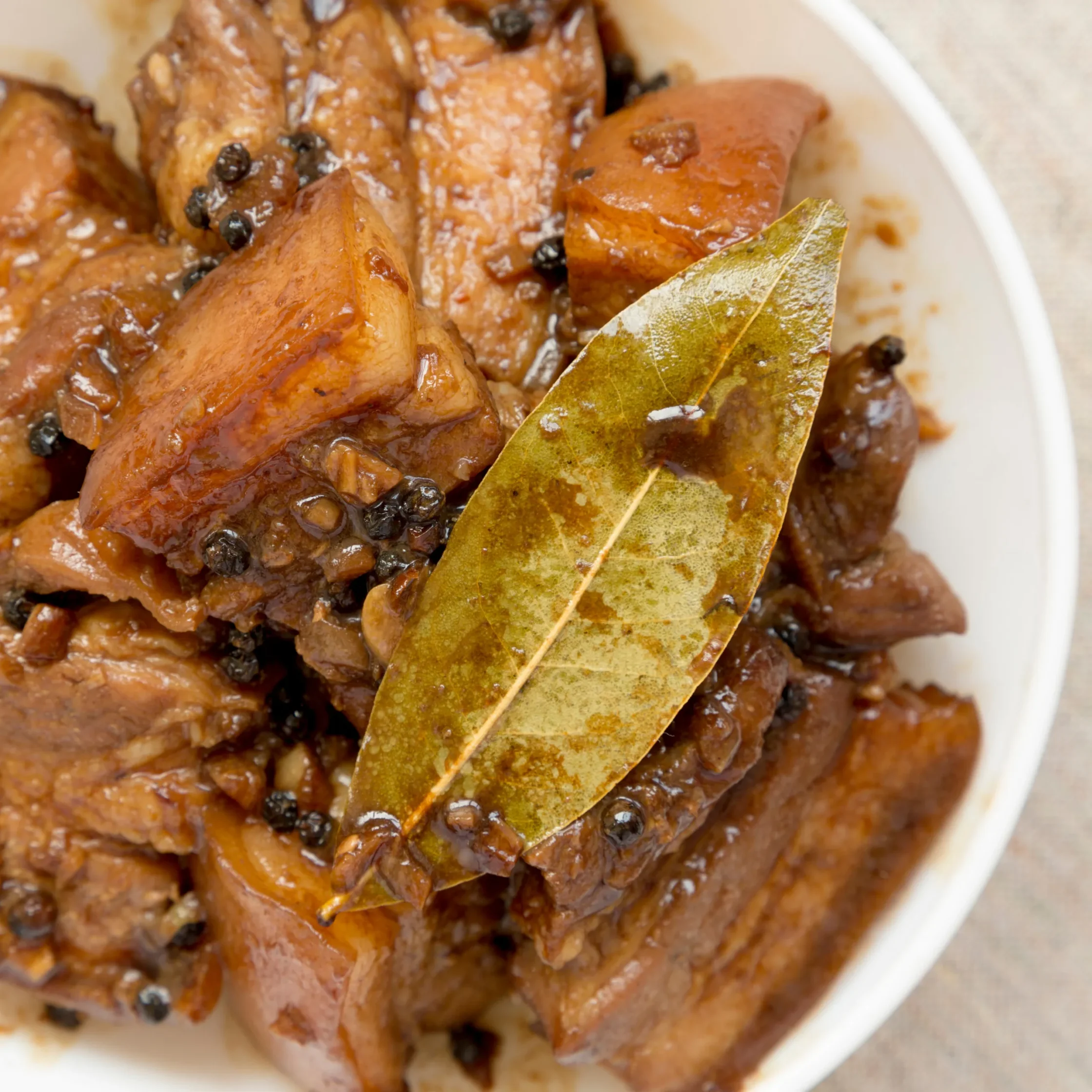 Close-up of glazed roasted meat with a bay leaf and scattered black peppercorns in sauce, on a white plate.